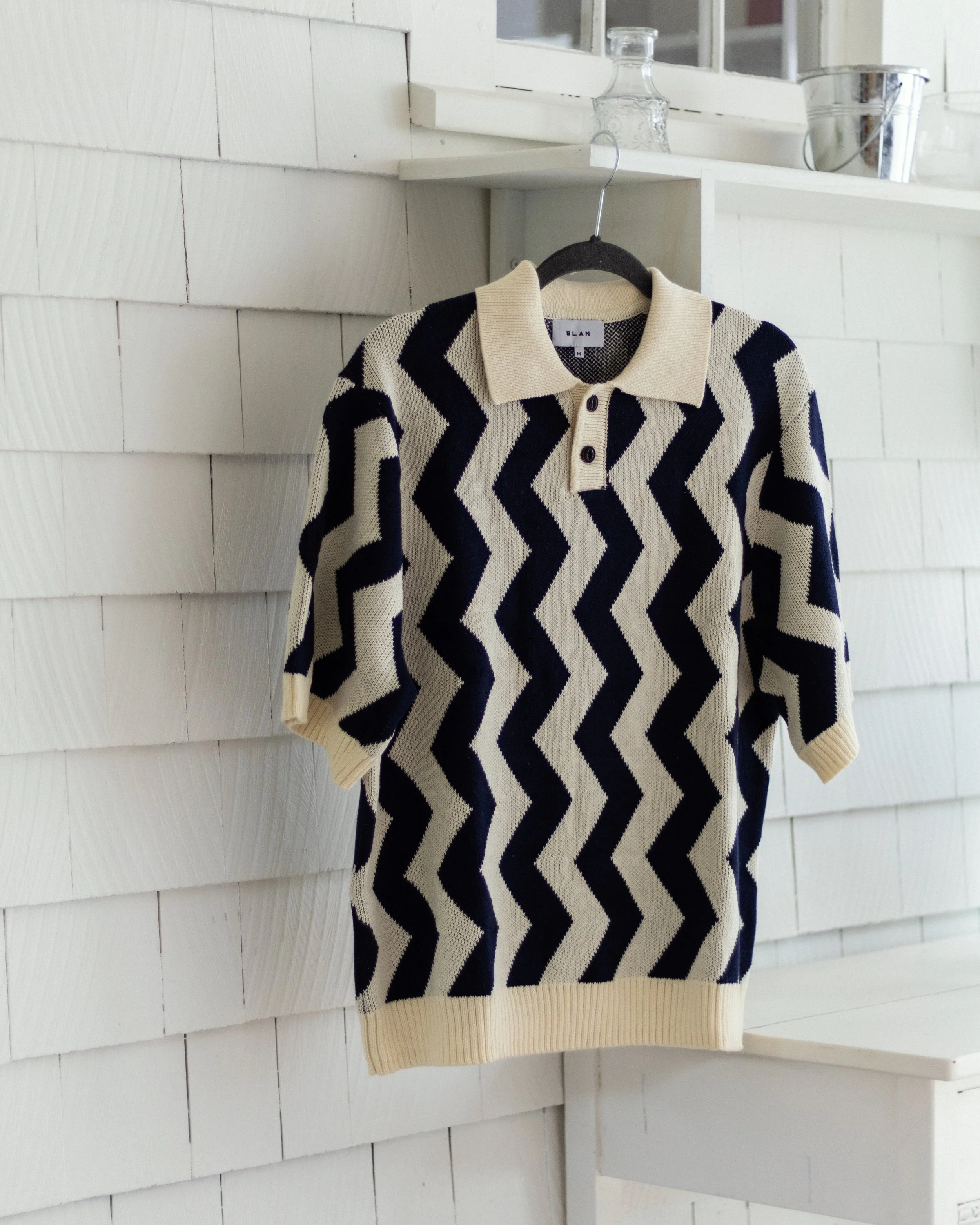 A cream-colored sweater with black zigzag pattern hanging on a black hanger. The sweater has a collar and buttons at the top, and is displayed against a white wooden wall with a shelf above, holding a glass bottle and a metal bucket.