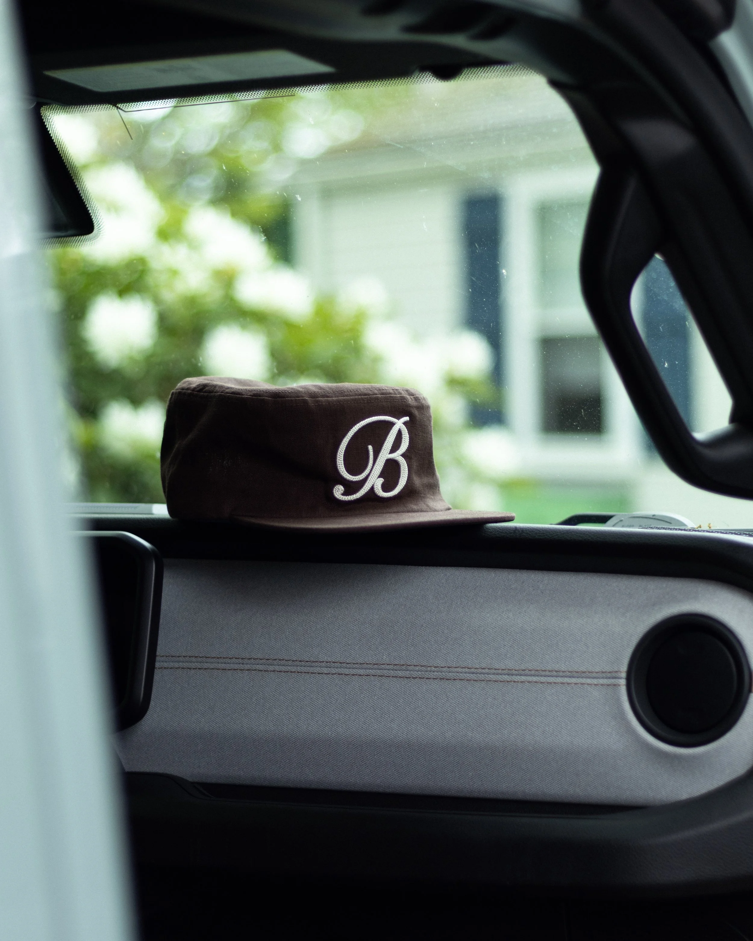 A brown cap with a white embroidered letter B on the front sitting on a car dashboard, viewed through the windshield with blurred greenery and house in the background.