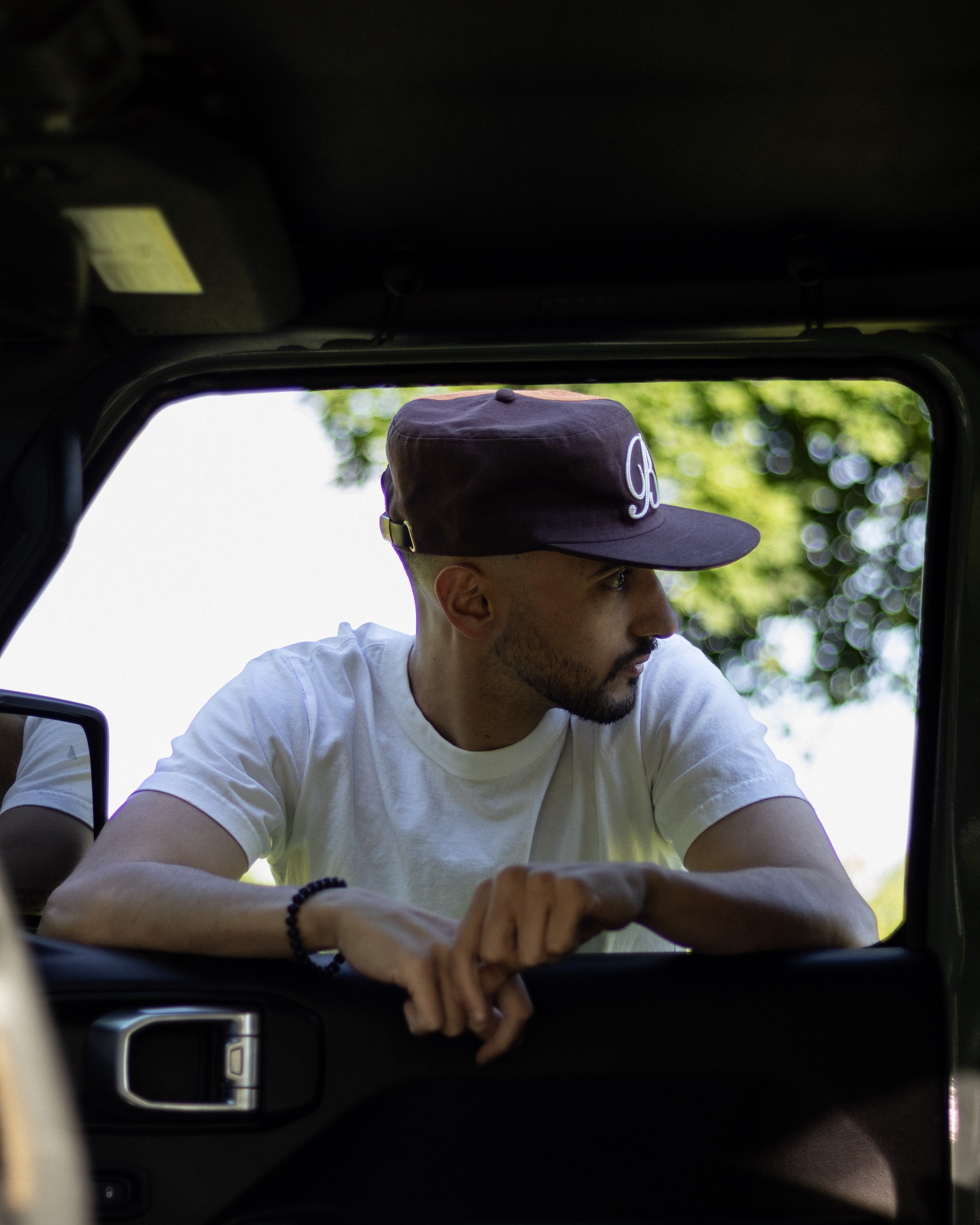 A man with a beard and sunglasses wearing a black cap and white T-shirt looking downward, seen from inside a car through the open window with blurred trees in the background.