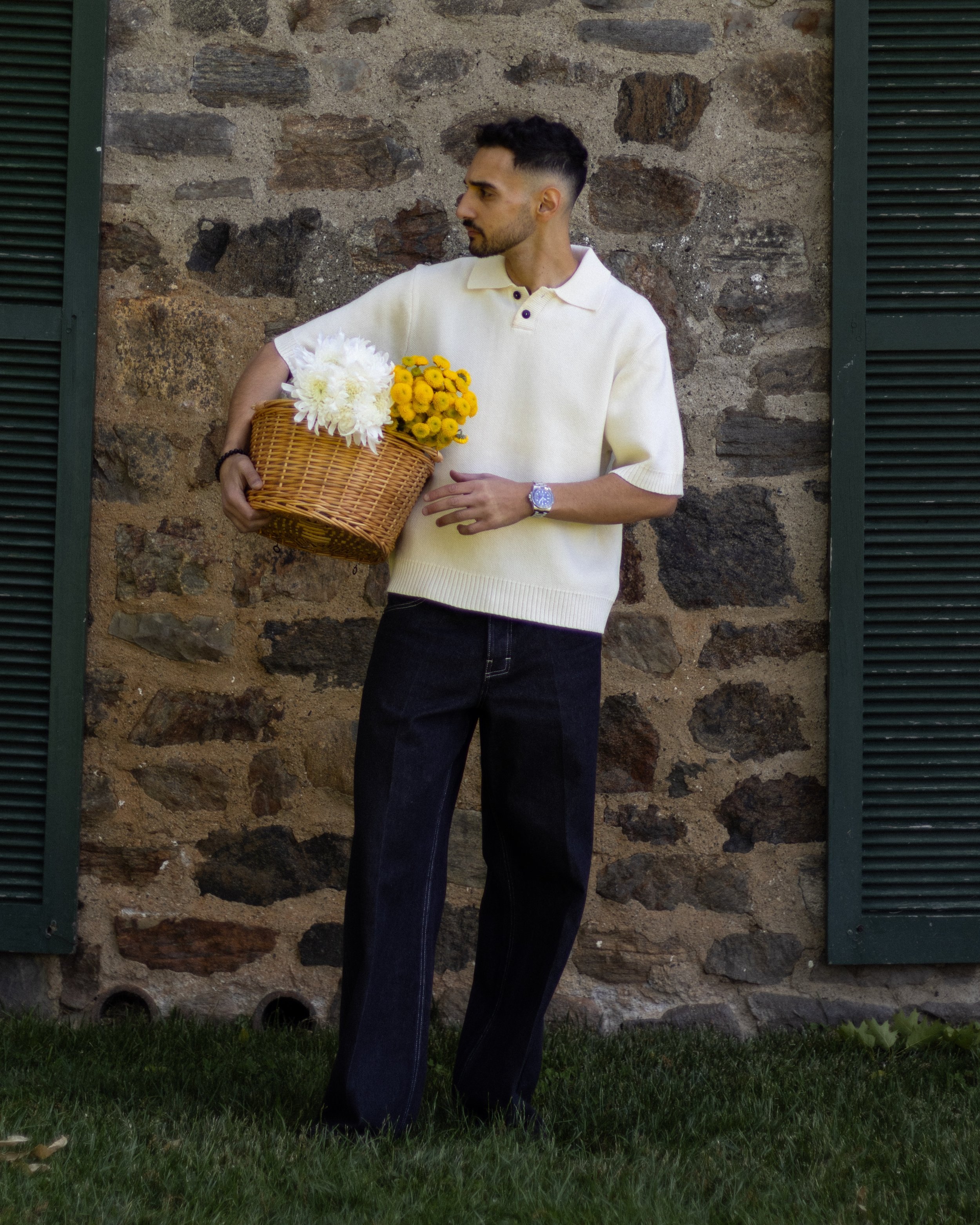 A man with short dark hair and a beard holding a wicker basket filled with white and yellow flowers stands against a rustic stone wall with green window shutters. He is wearing a cream-colored short-sleeve sweater and dark jeans, looking to his left.