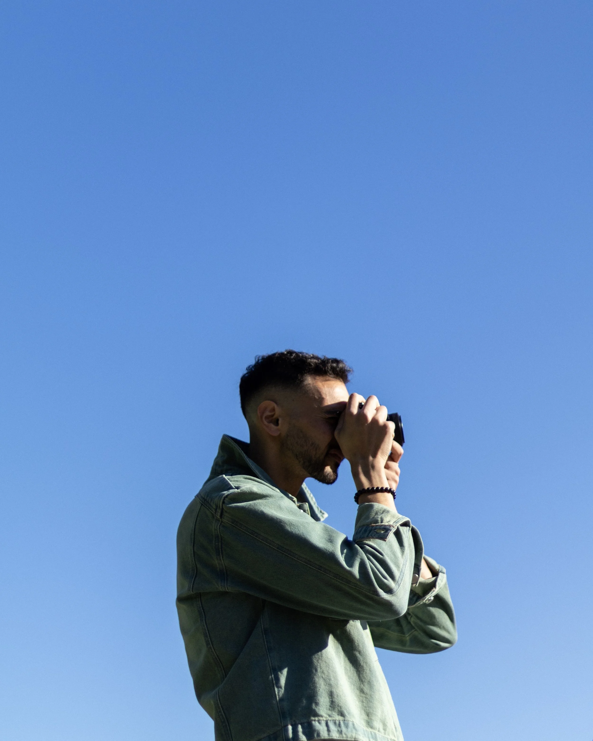 A young man with dark hair and a beard is standing outdoors against a clear blue sky, looking through binoculars.