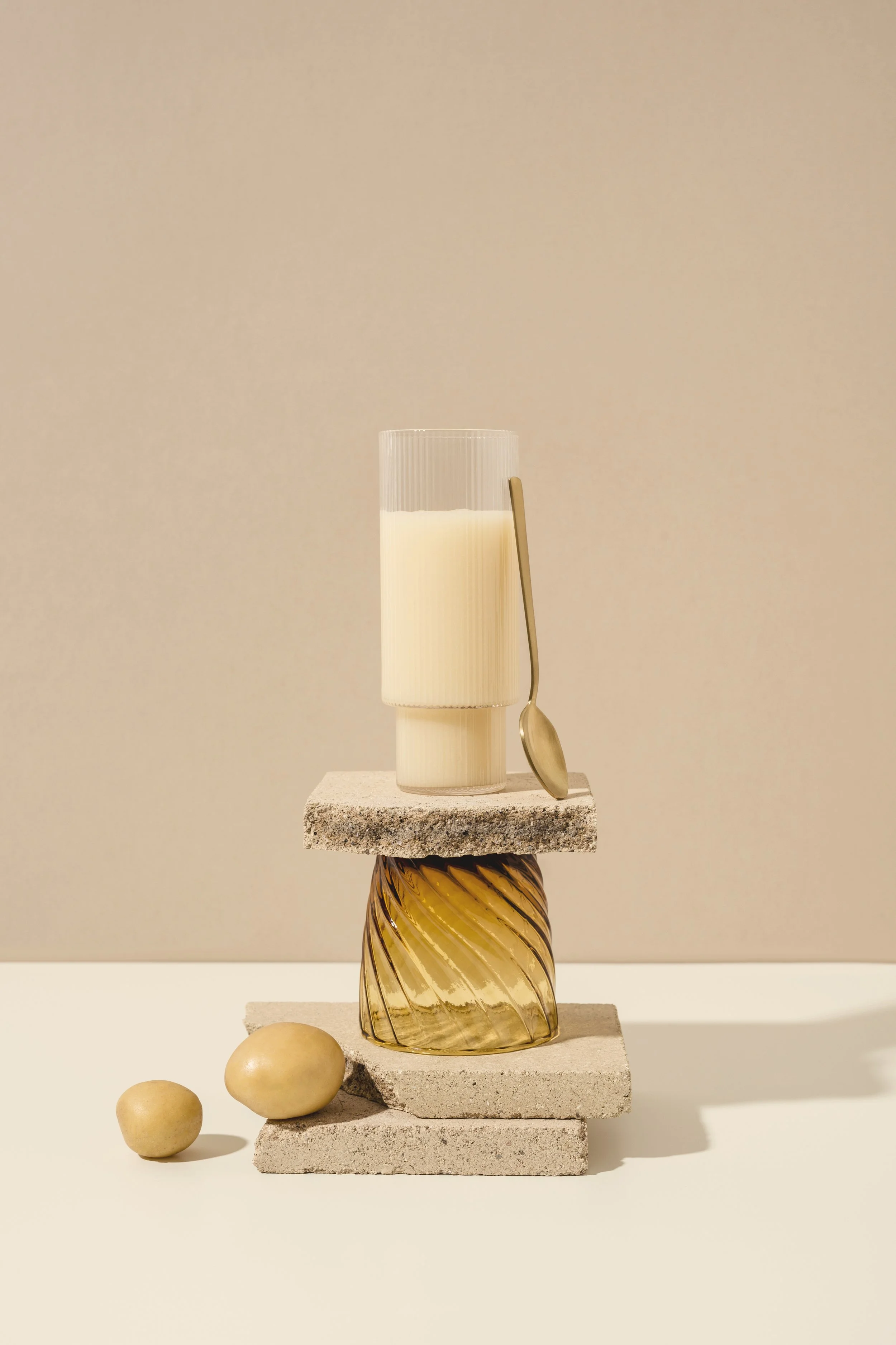 Una composizione artistica con vari oggetti, tra cui un bicchiere di latte, una colonna di pietra e vetro, alcune pietre e un cucchiaino, disposti in modo equilibrato.