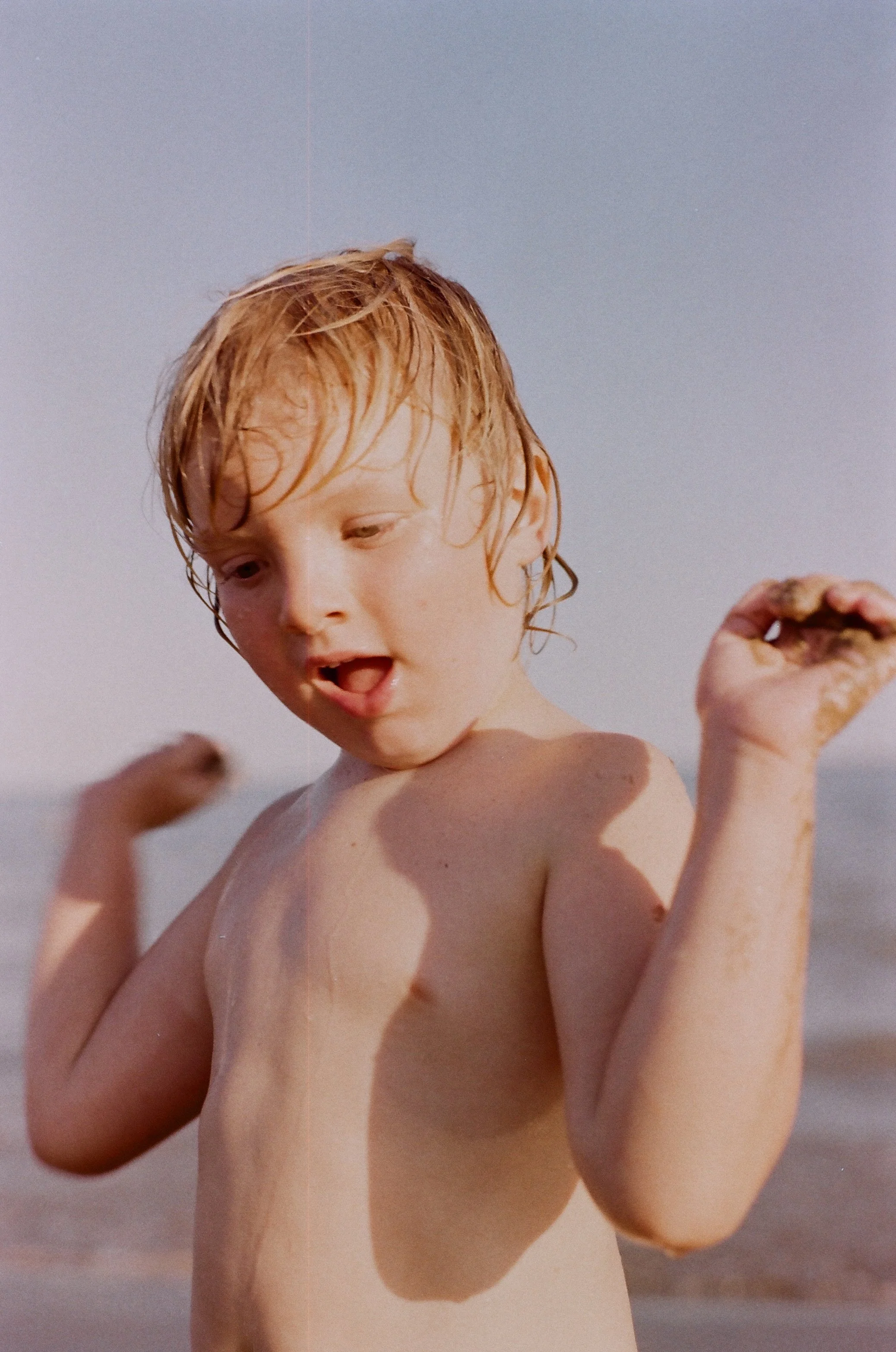 Junge am Strand, mit nassblondem Haare, die Freibad- oder Strandkleidung trägt, an den Schultern und Armen mit Sand. Im Hintergrund das Meer, sonniges Wetter.