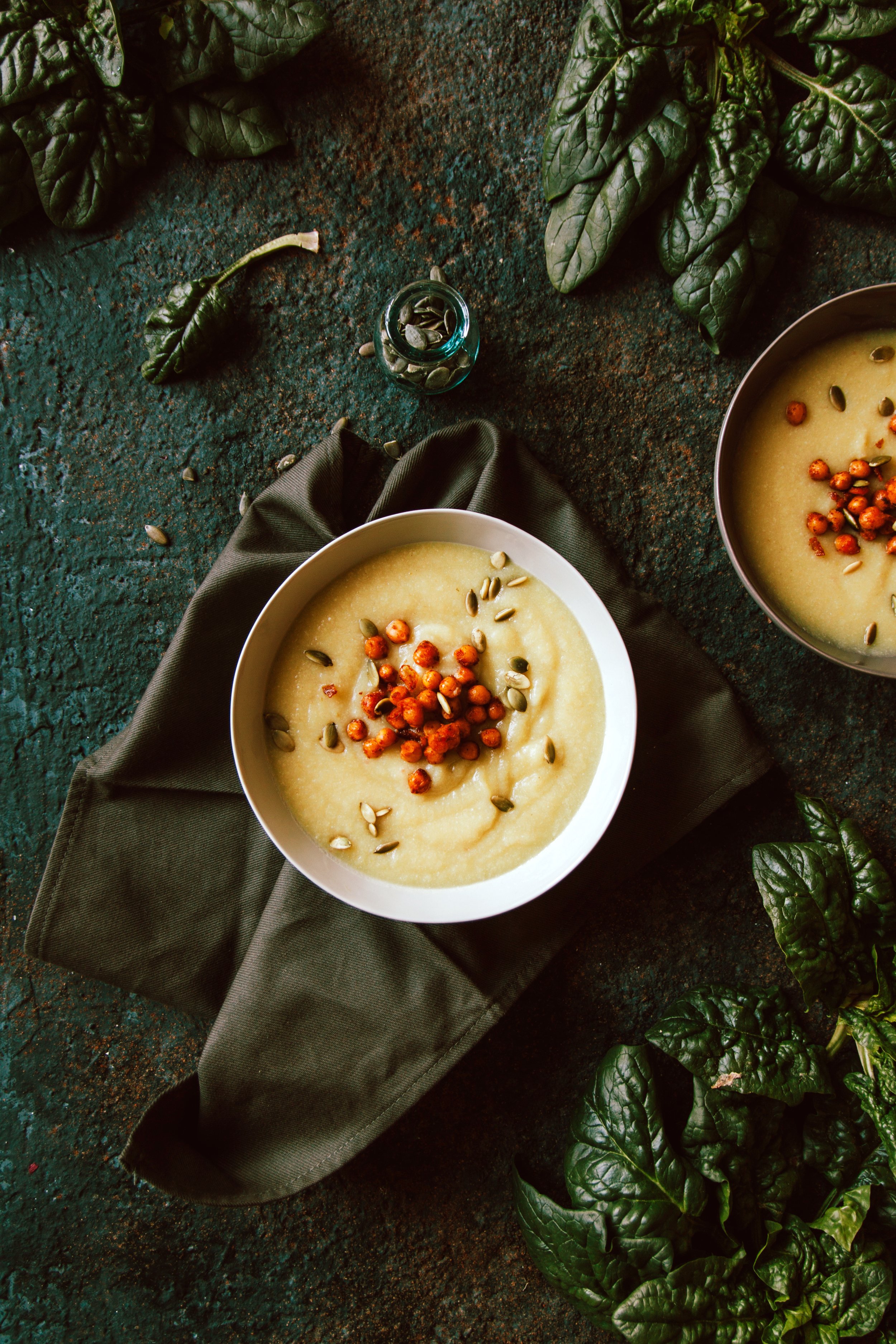 Une soupe crémeuse garnie de graines de courge et de baies roses. Sur un fond vert texturé avec des feuilles de basilic et un petit flacon de graines.