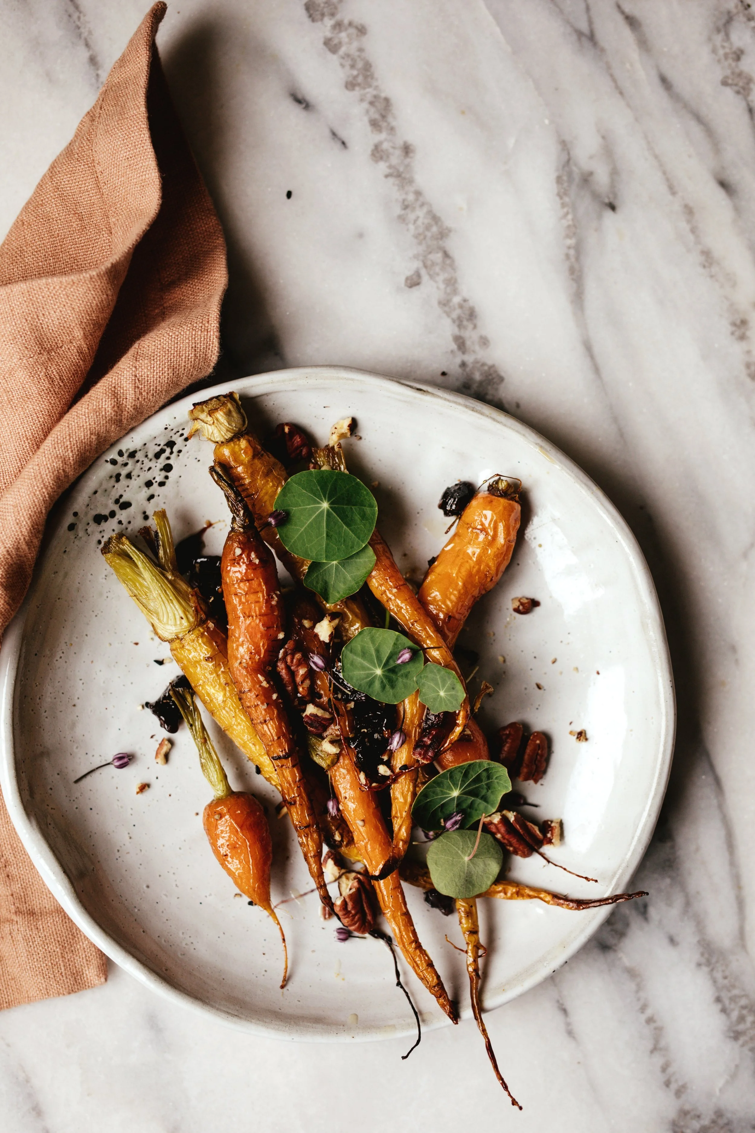 Carottes rôties présentées dans une assiette blanche, garnies de feuilles de lierre et de noix, sur une surface en marbre avec un torchon en tissu beige à gauche.