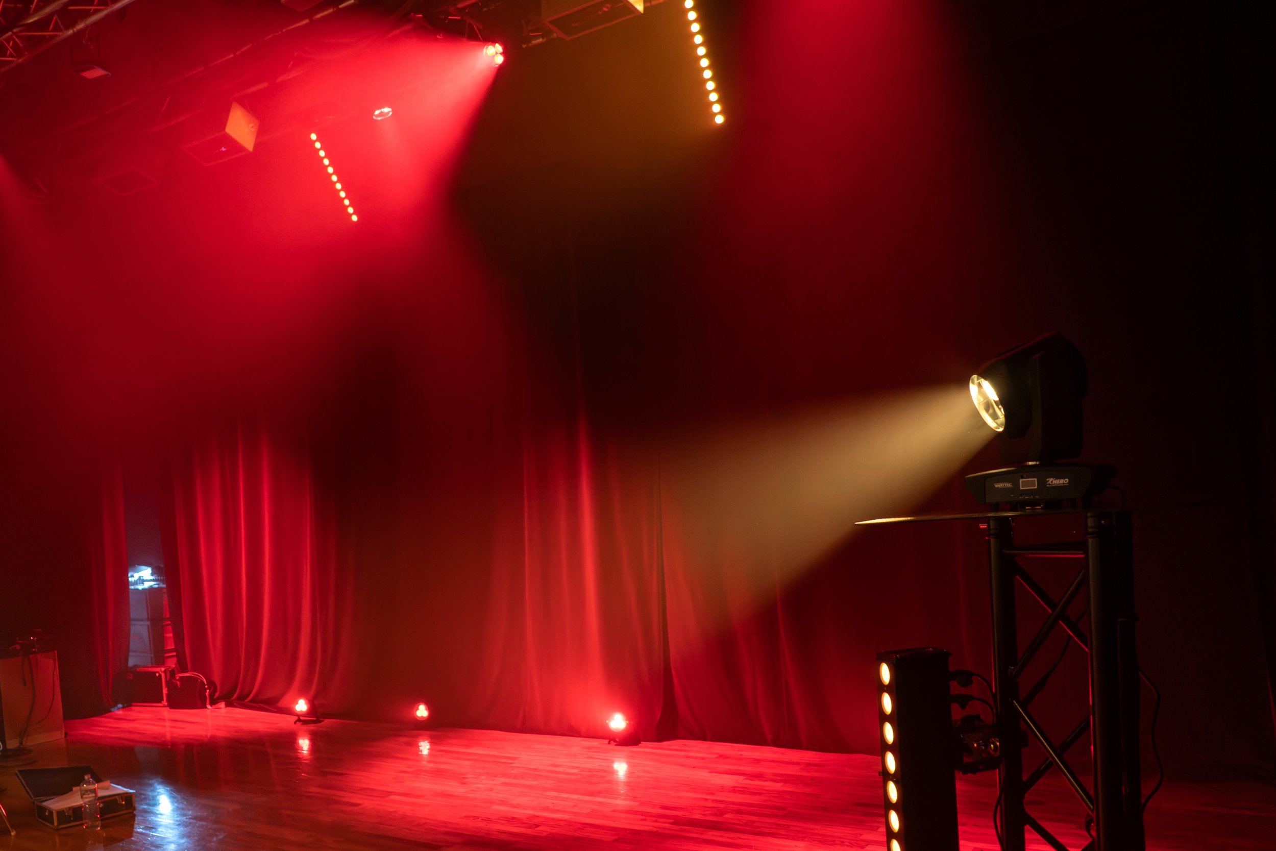Scène de théâtre ou de concert avec des lumières rouges et une scène vide, des rideaux rouges en arrière-plan, un projecteur éclairant la scène, et des équipements techniques sur le côté.