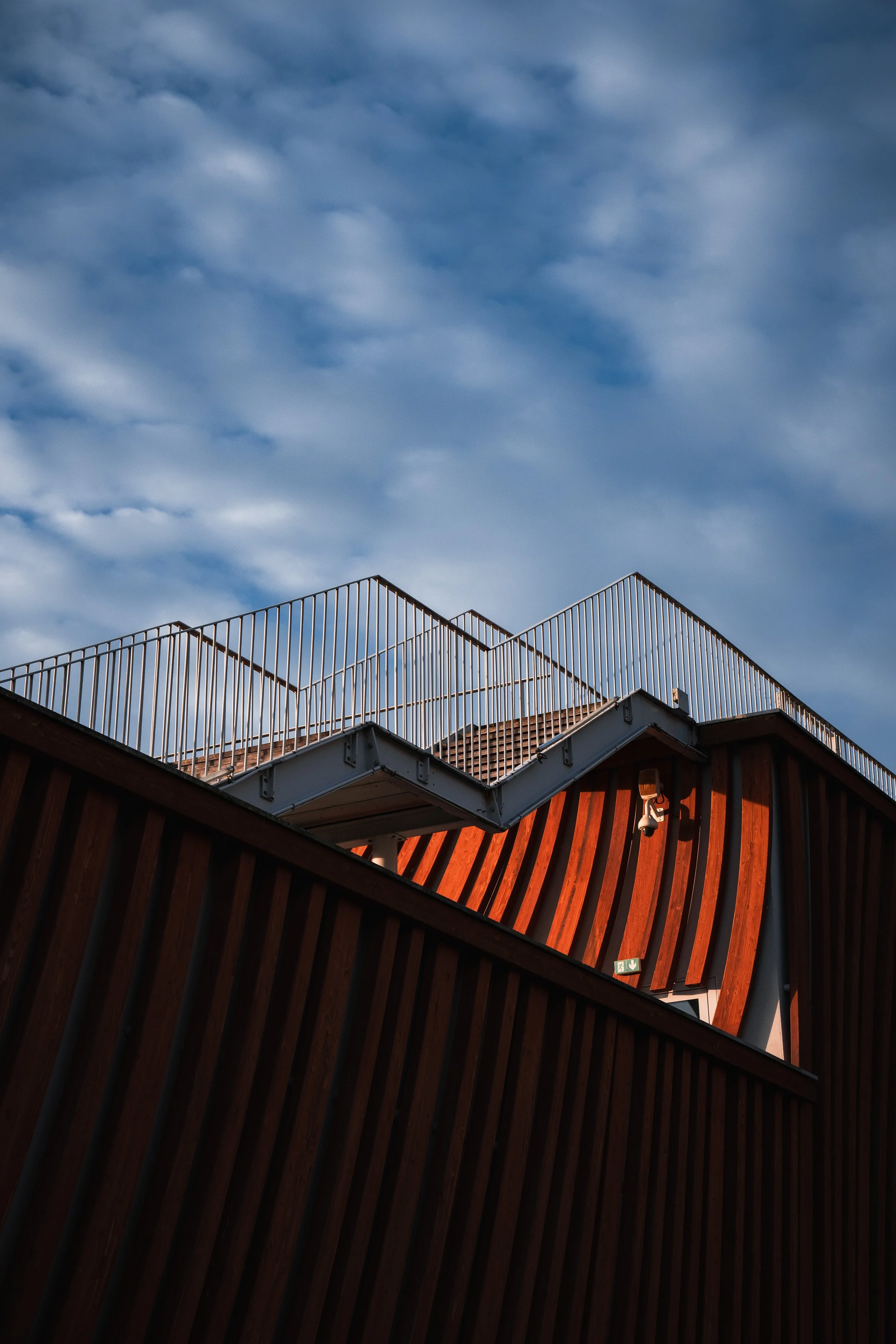 Vue d'un bâtiment moderne avec des structures en bois courbes et des balustrades en métal sous un ciel nuageux.