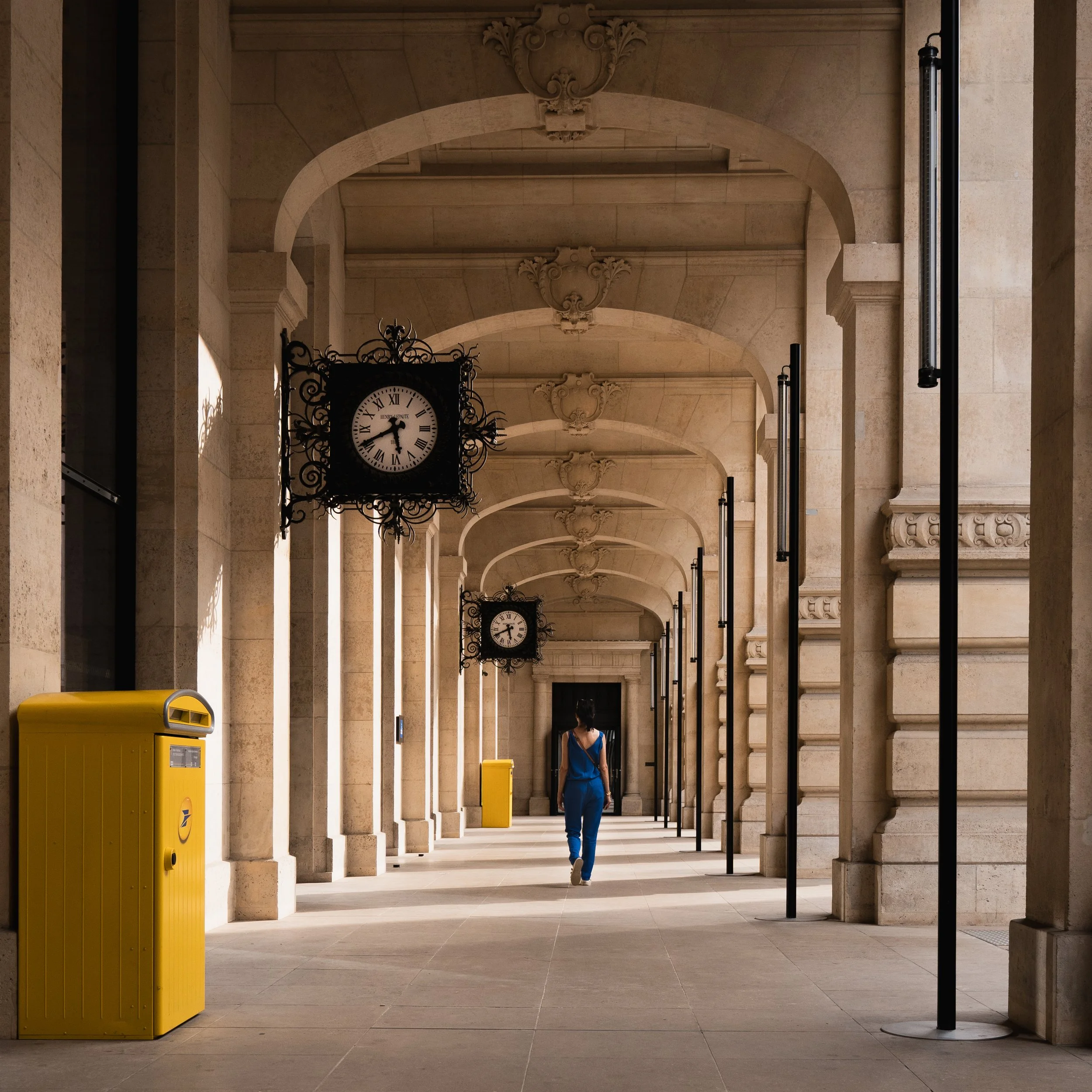 Un couloir architectural avec des horloges murales en fer forgé, des colonnes en pierre, un sol en pierre claire, une femme marchant en jeans bleus, et une boîte aux lettres jaune à gauche.