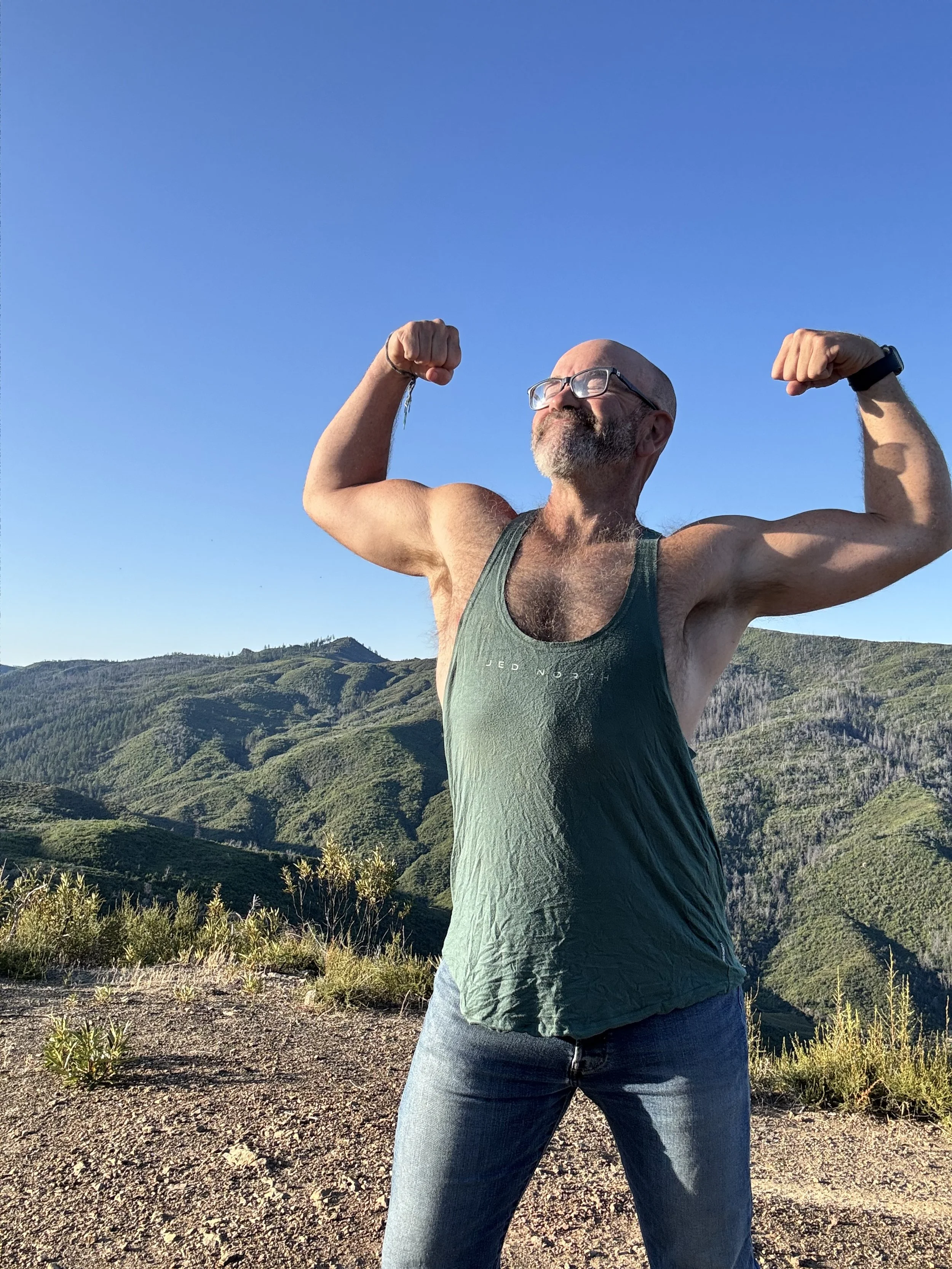 A bearded man wearing glasses and a green tank top flexing his muscles outdoors on a mountain trail with green hills and blue sky in the background.