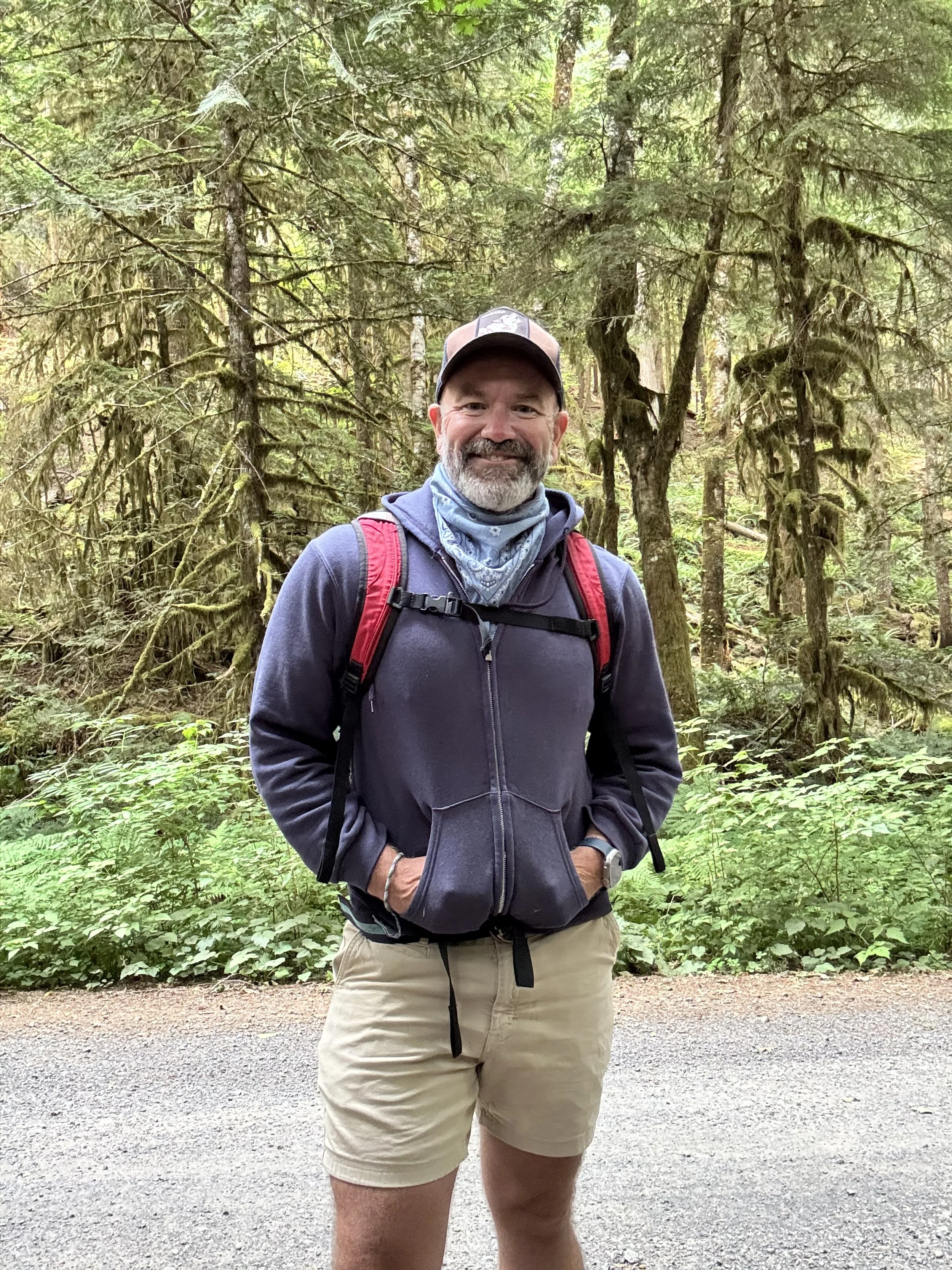 A man with a beard, wearing a baseball cap, gray hoodie, and tan shorts, stands on a trail in a forest with dense green trees and foliage, smiling at the camera.