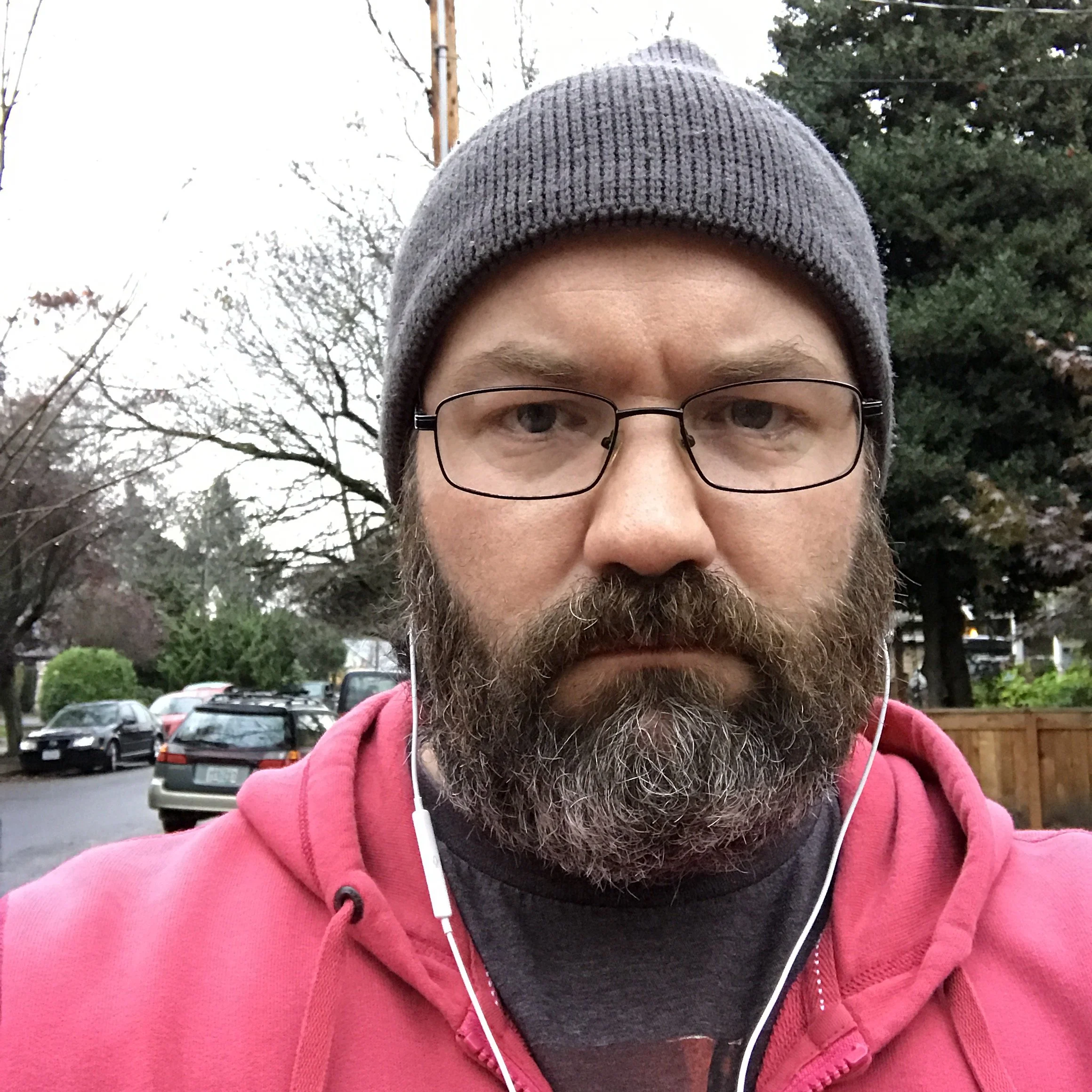 A man with glasses and a beard wearing a gray beanie and pink hoodie taking a selfie outdoors in a residential area with trees and parked cars in the background.