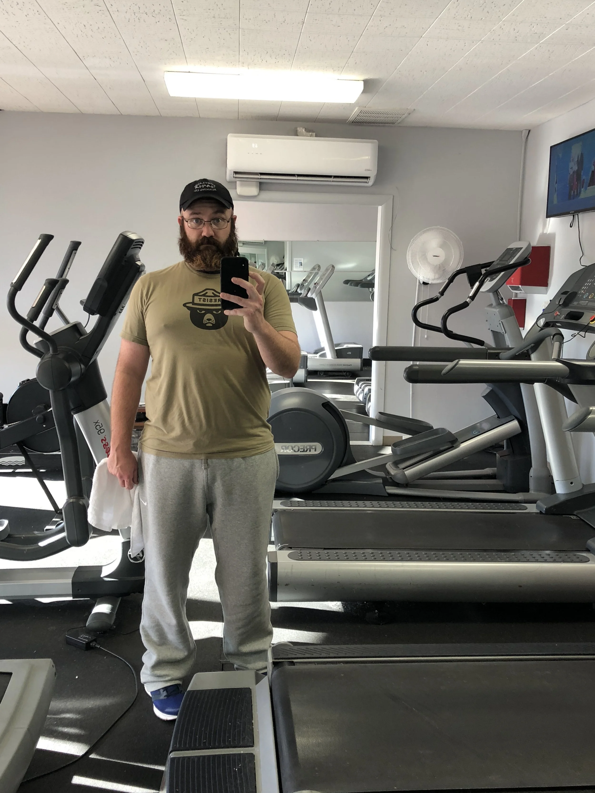 A man with glasses and a beard taking a mirror selfie in a gym. The man is wearing a baseball cap, a beige T-shirt with a graphic, and gray sweatpants. The gym has exercise equipment, including treadmills and ellipticals, with a TV mounted on the wall in the background.