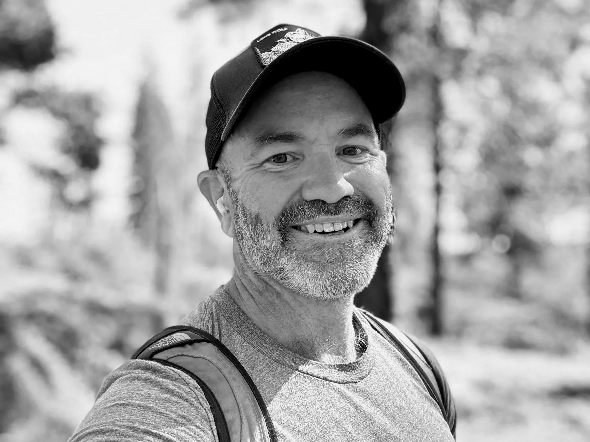 A smiling man in a baseball cap outdoors, with a backpack, in a blurred forest background.