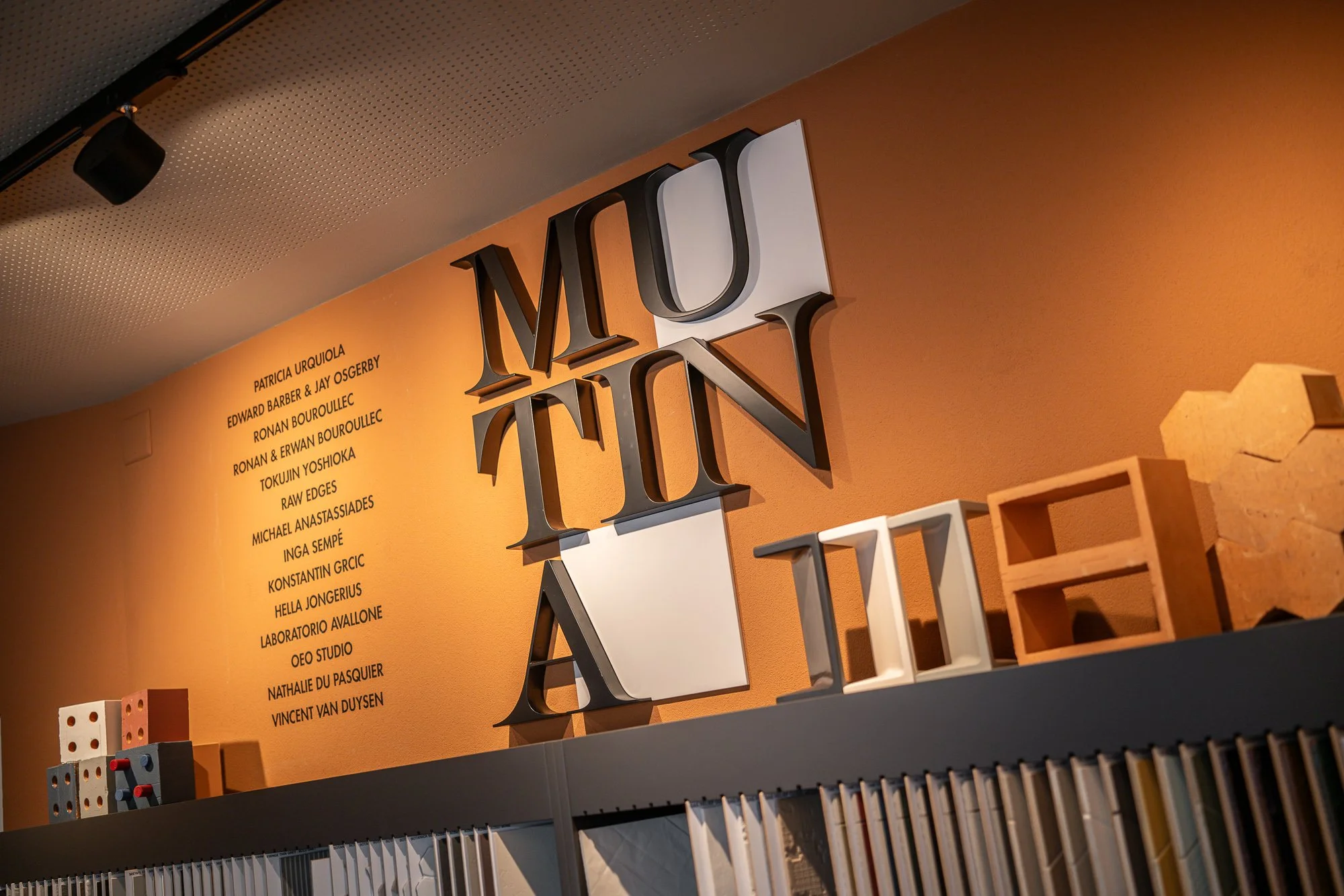 Wand mit der Aufschrift 'MUTINA', daneben eine Liste von Namen, ein Regal mit Farben und Musterproben.