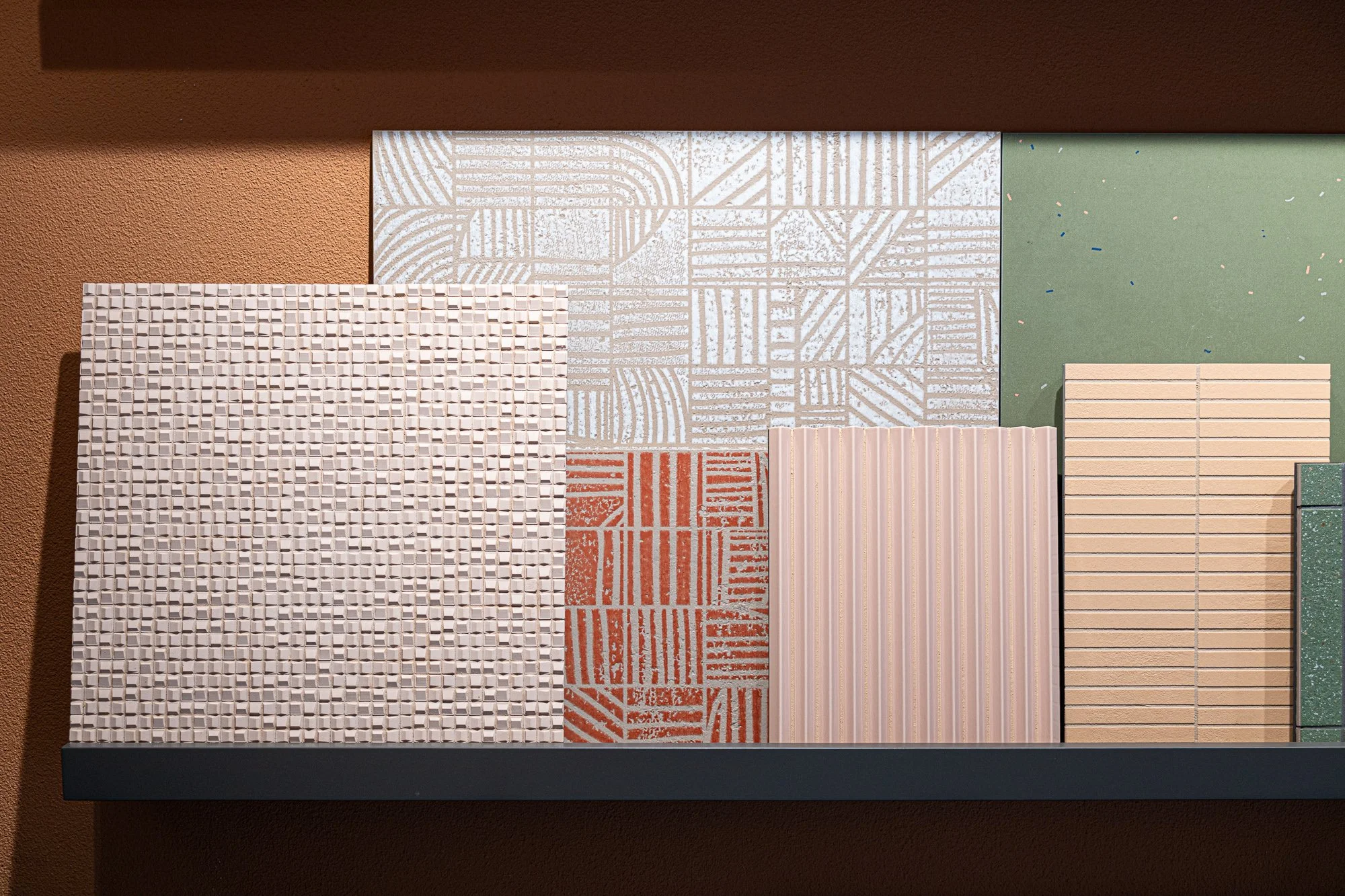 Verschiedene Fliesenmustern und -oberflächen auf einer schwarzen Regalplatte vor einem grünen und terrakottafarbenen Hintergrund.