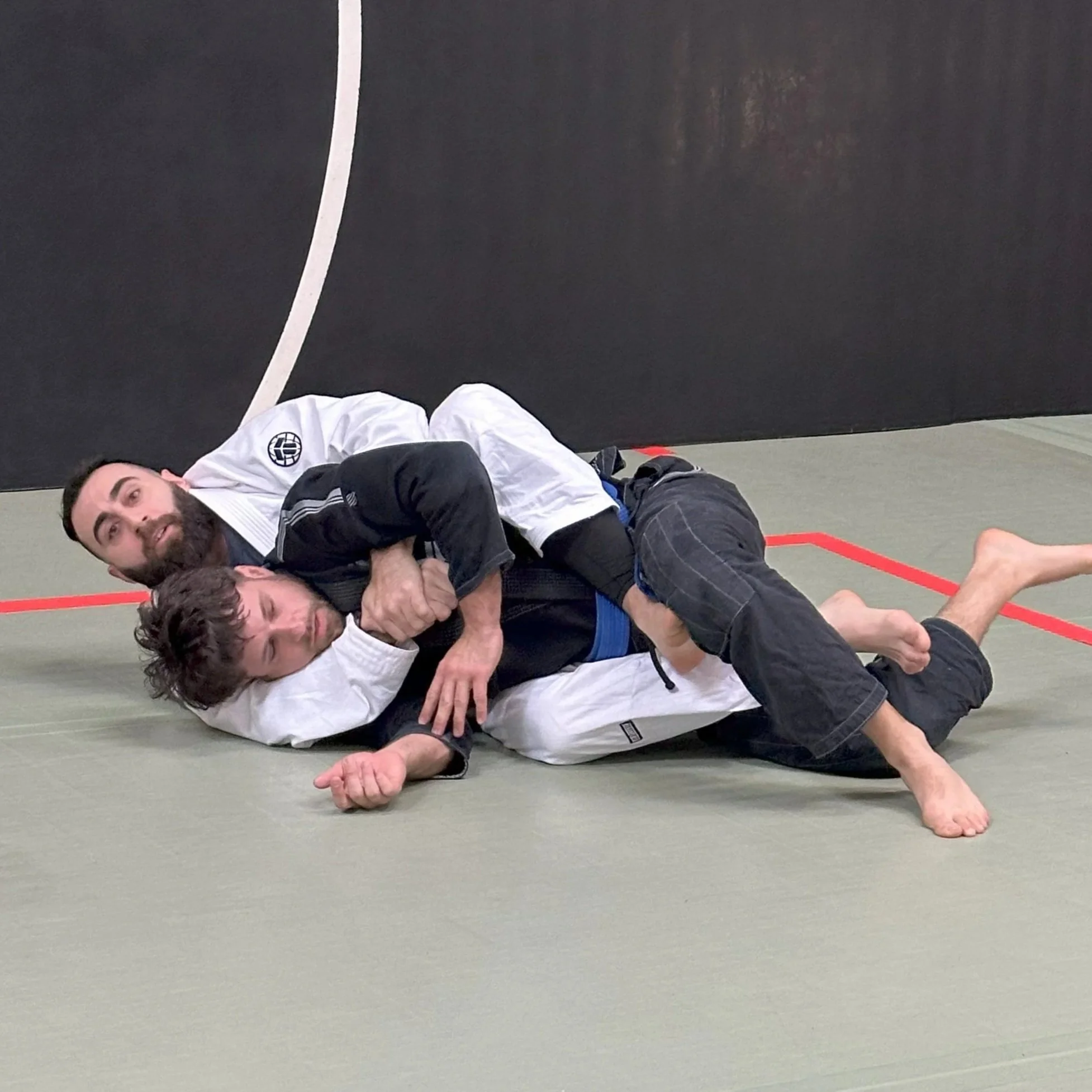 Two men are practicing Brazilian Jiu-Jitsu on a mat, one in a white gi and the other in a black gi, engaged in a grappling move.