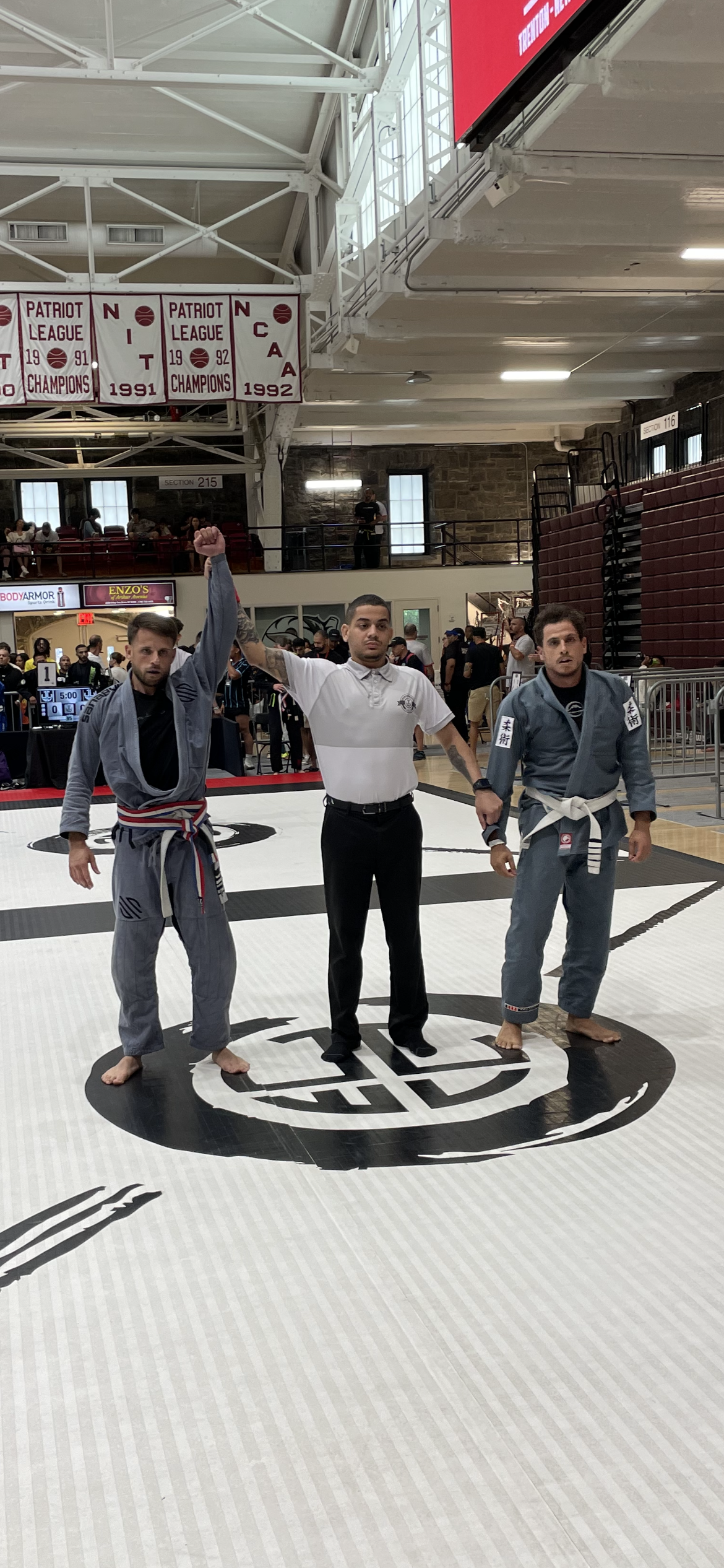 A Brazilian Jiu-Jitsu competition with two competitors in Gis and a referee in the center raising one competitor's arm in victory. The venue is an indoor sports arena with banners displaying championship titles hanging from the ceiling and spectators in the background.