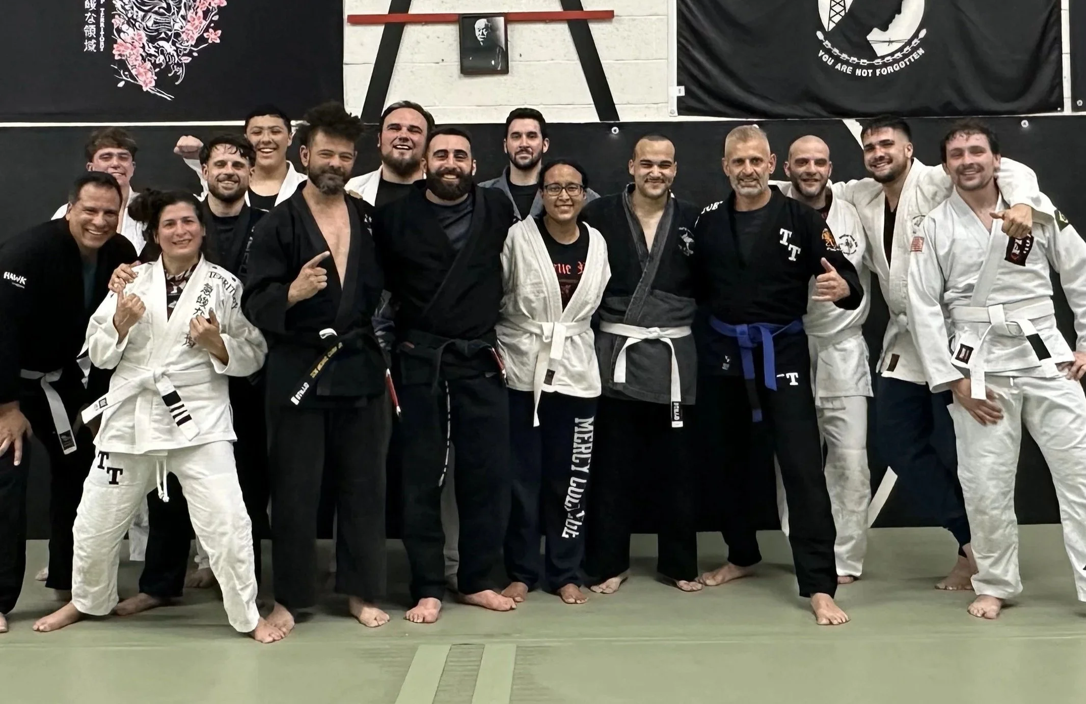 A group of martial arts practitioners, some in white and others in black uniforms, standing together on a training mat in a dojo, smiling and posing for a photo.