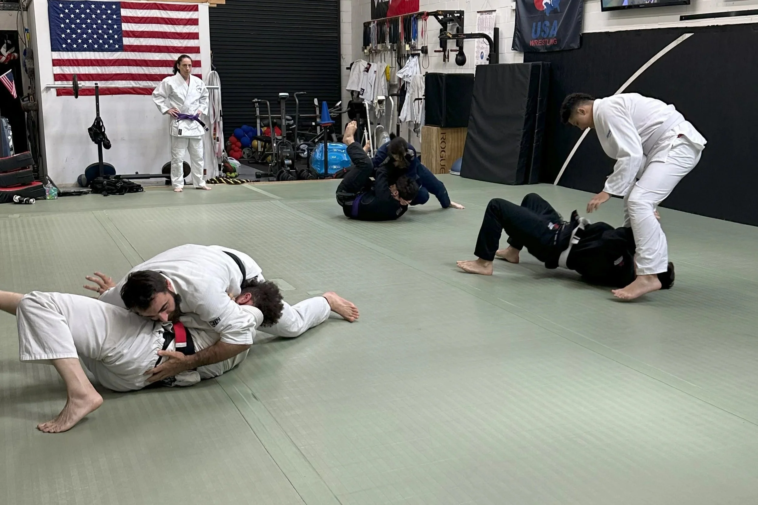 Martial arts class in a gym with students practicing Brazilian Jiu-Jitsu techniques, some on the mat and others standing or observing.