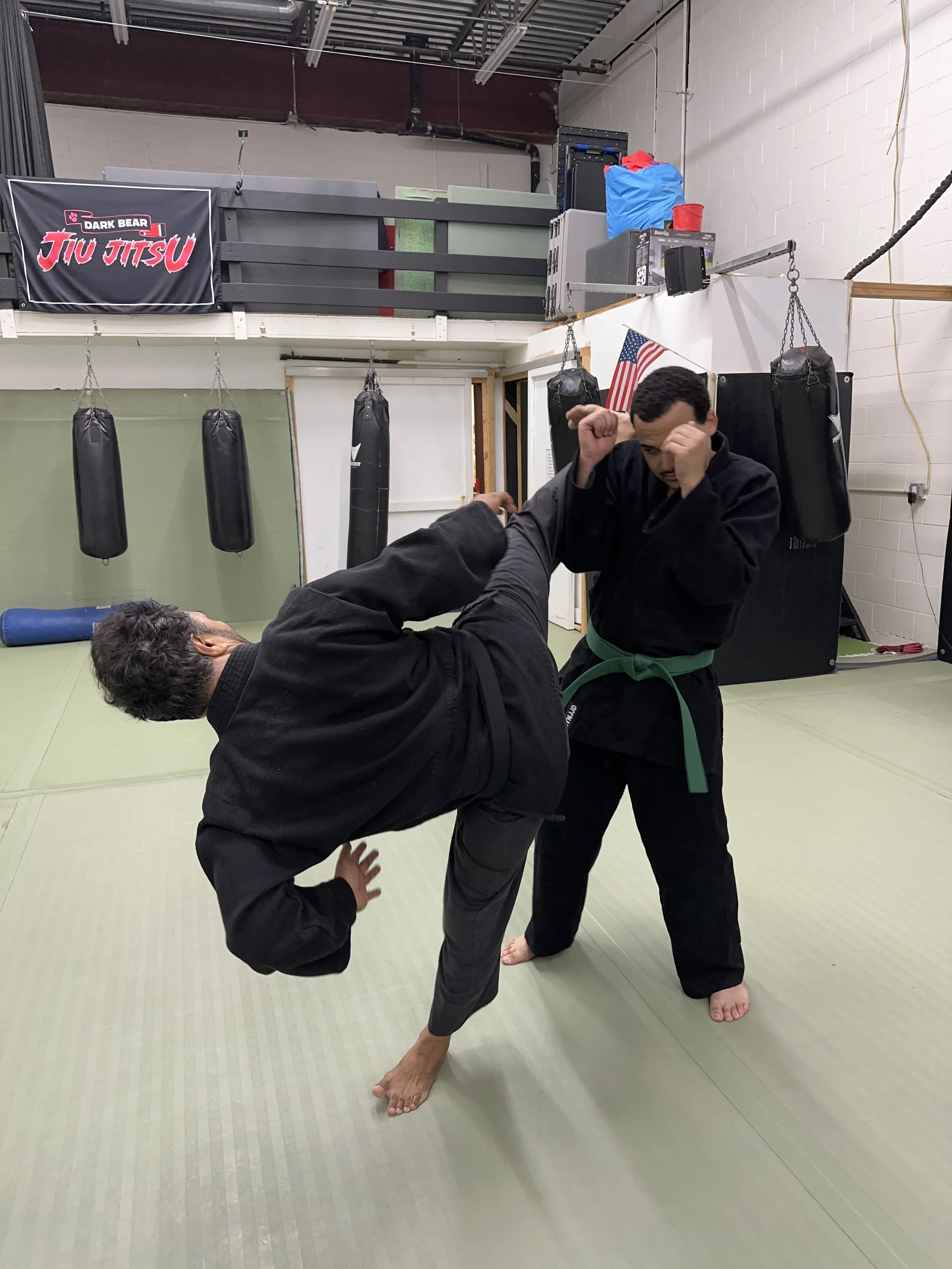 Two men practicing Brazilian Jiu-Jitsu on mats in a martial arts gym, one executing a throw on the other, with punching bags hanging in the background.