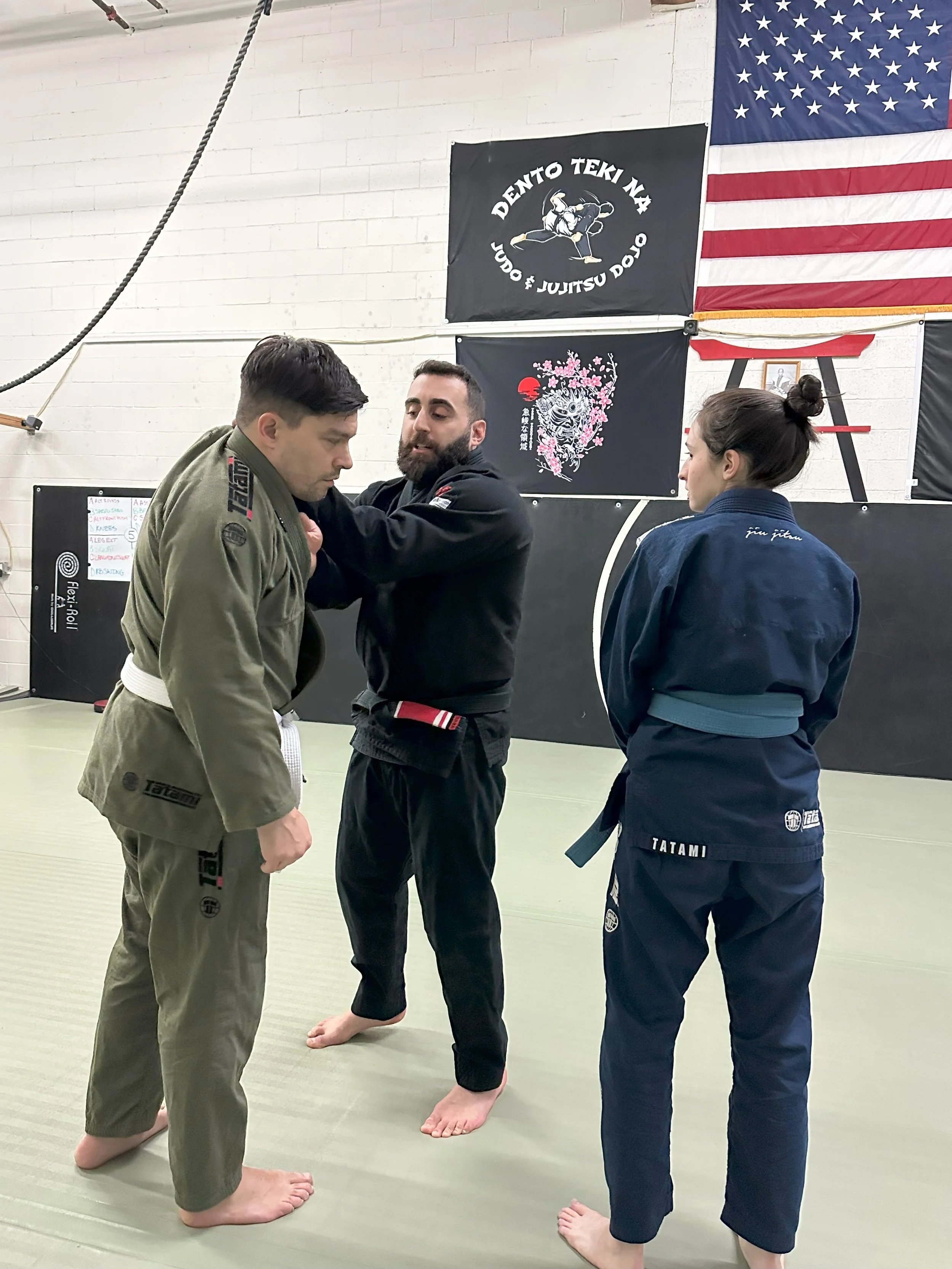 Two men practicing Brazilian Jiu-Jitsu with a woman observing in a martial arts gym. One man is wearing a green gi and the other a black gi, while the woman is wearing a dark blue gi.