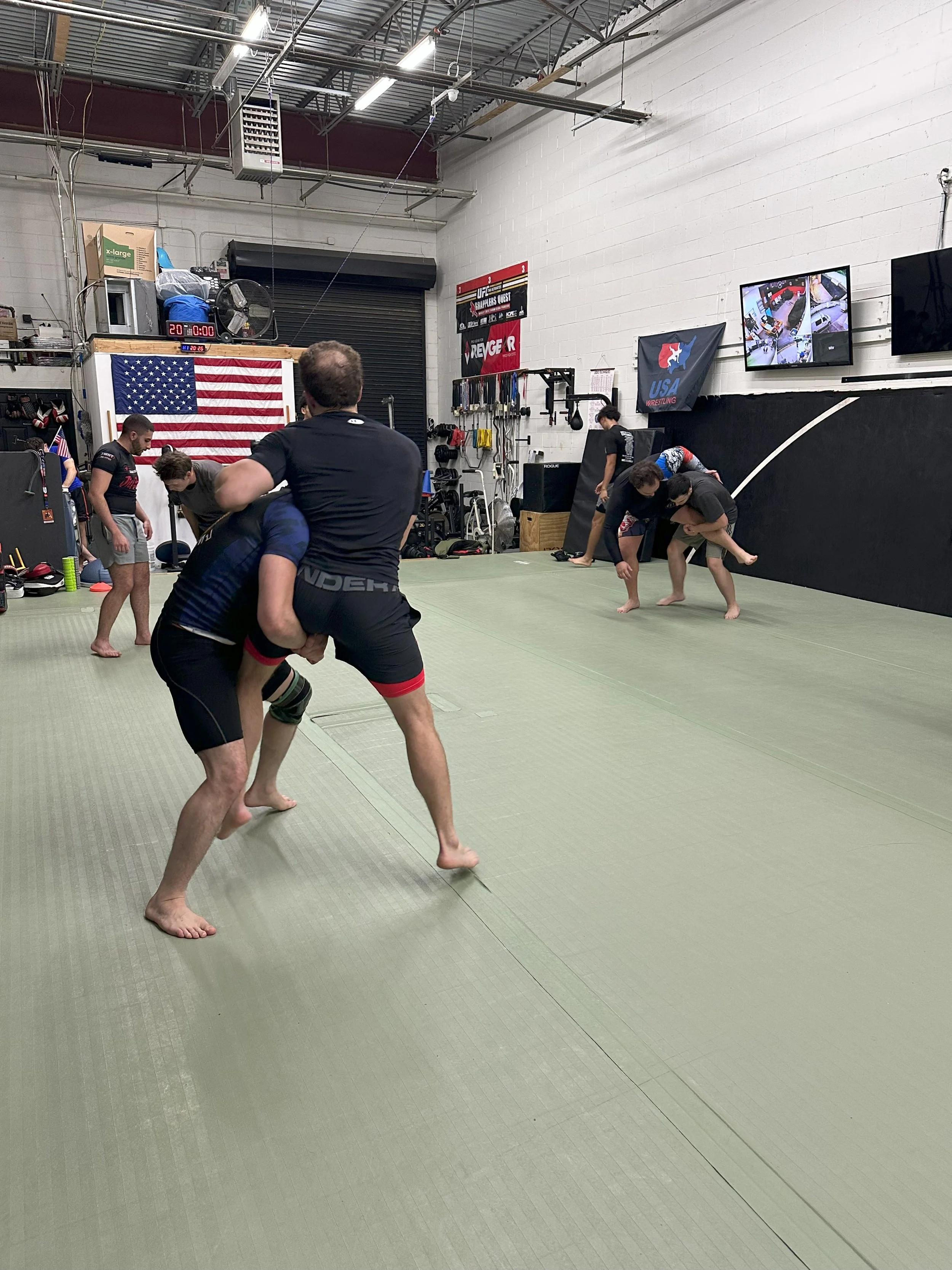 People practicing Brazilian Jiu-Jitsu on a green mat in a gym with American flags and wrestling banners on the walls.