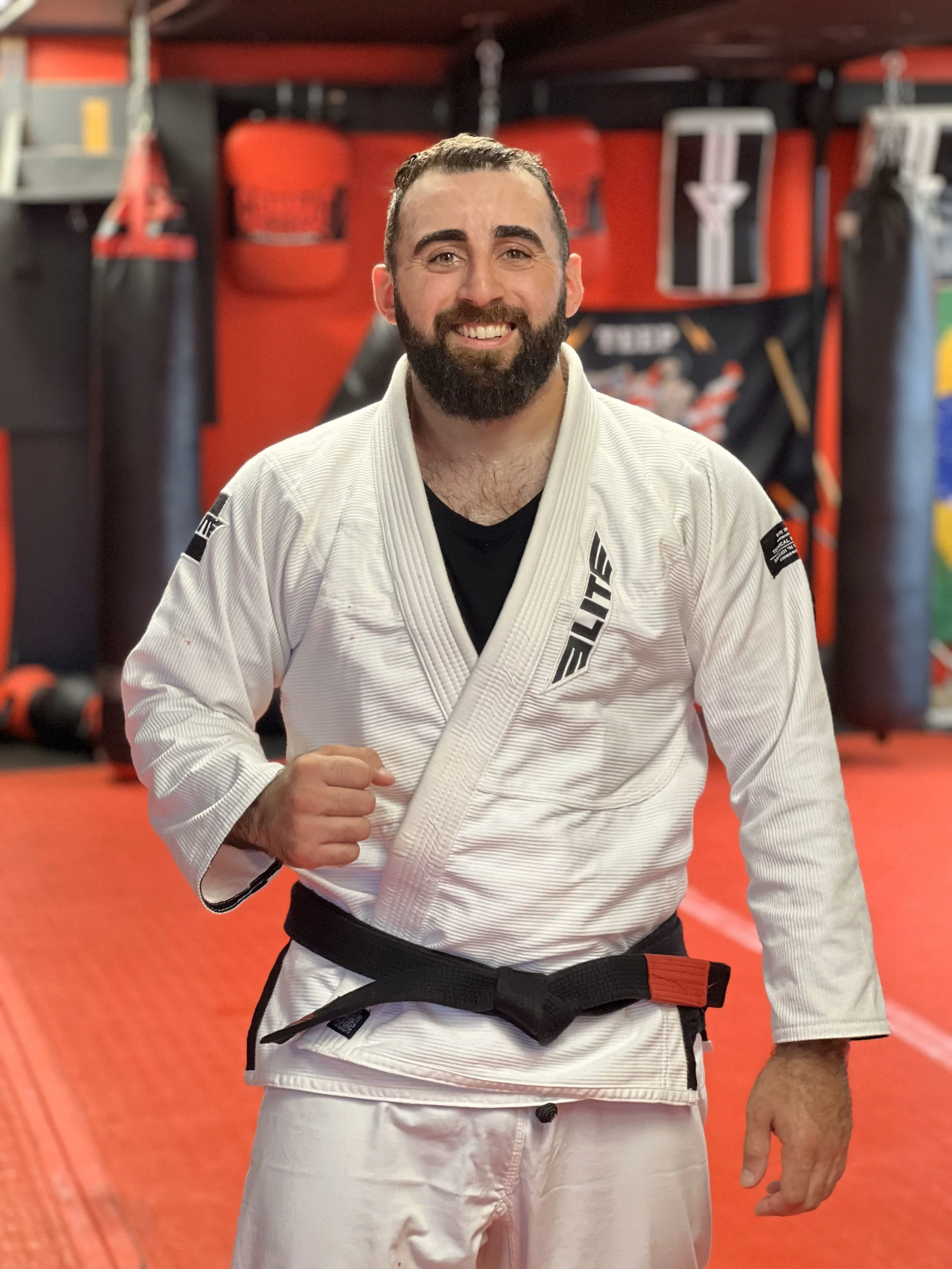 A smiling man in a white Brazilian Jiu-Jitsu gi with a black belt in a martial arts gym.