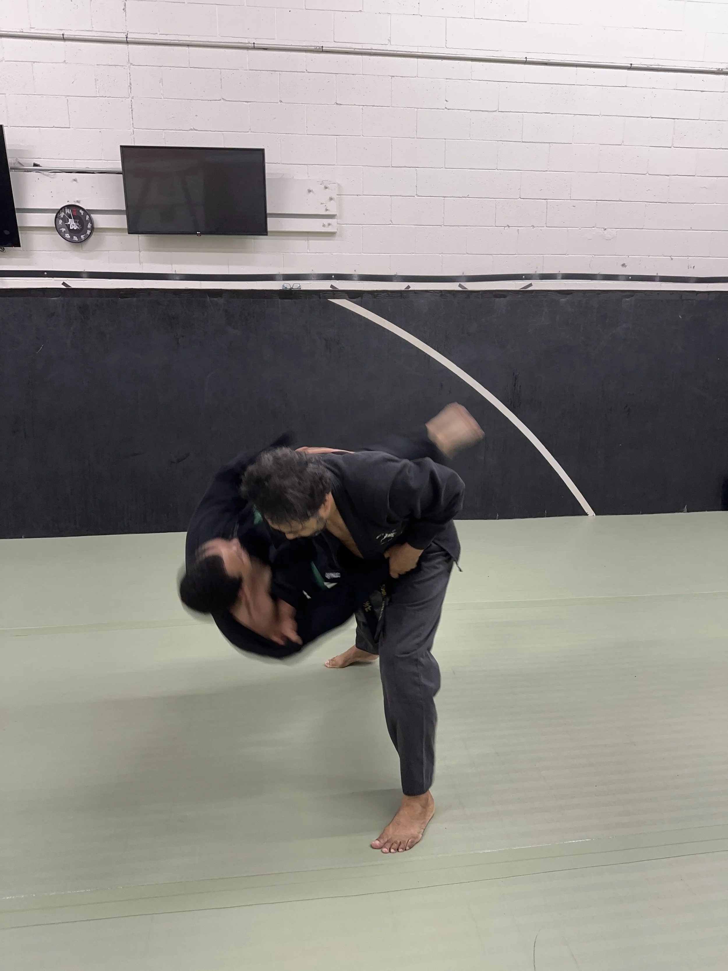 Two martial artists practicing Brazilian Jiu-Jitsu on a green mat in a training facility. One is executing a move while the other is defending.