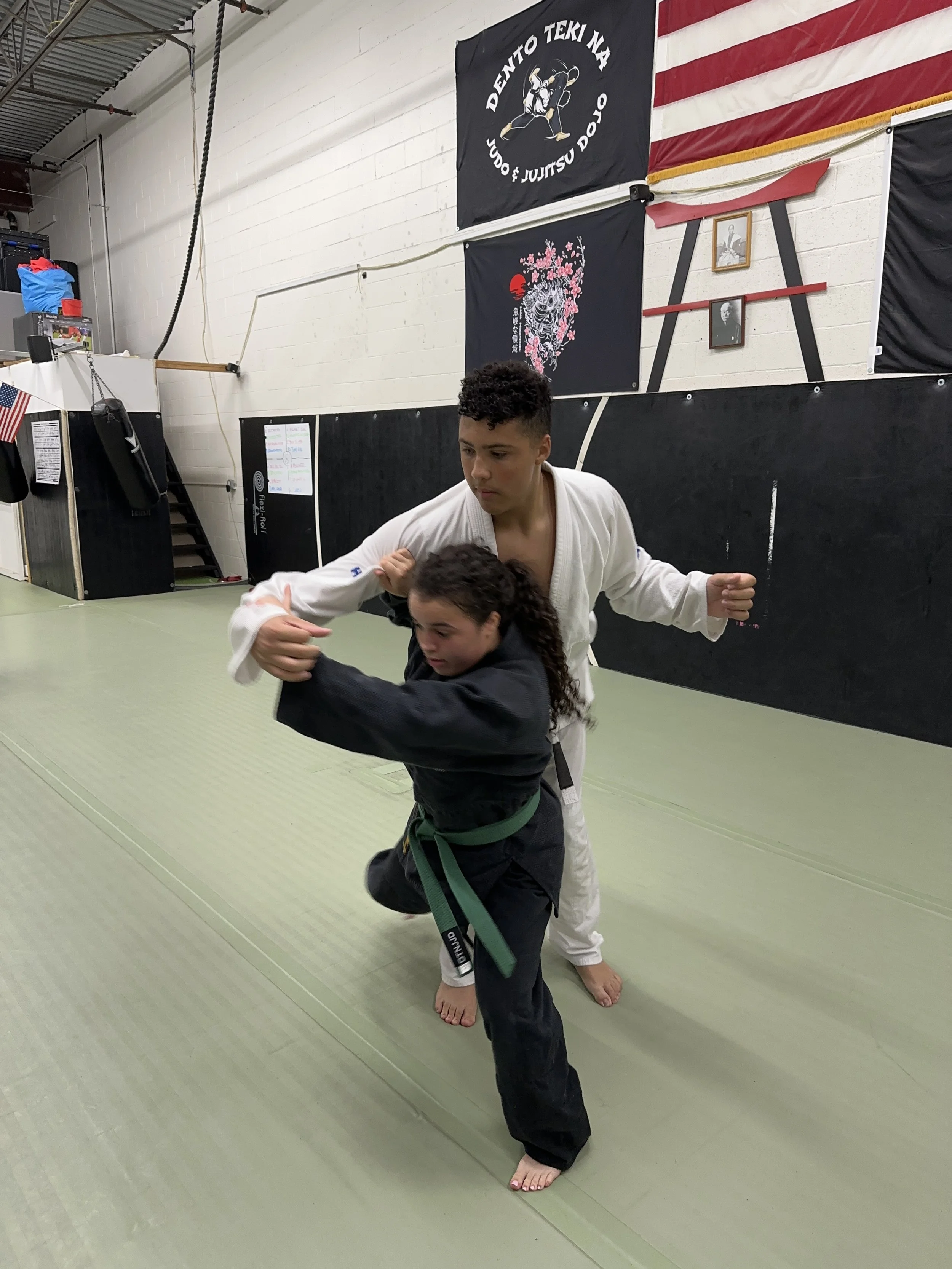 Two martial artists practicing judo in a dojo, with banners, flags, and photos on the wall.