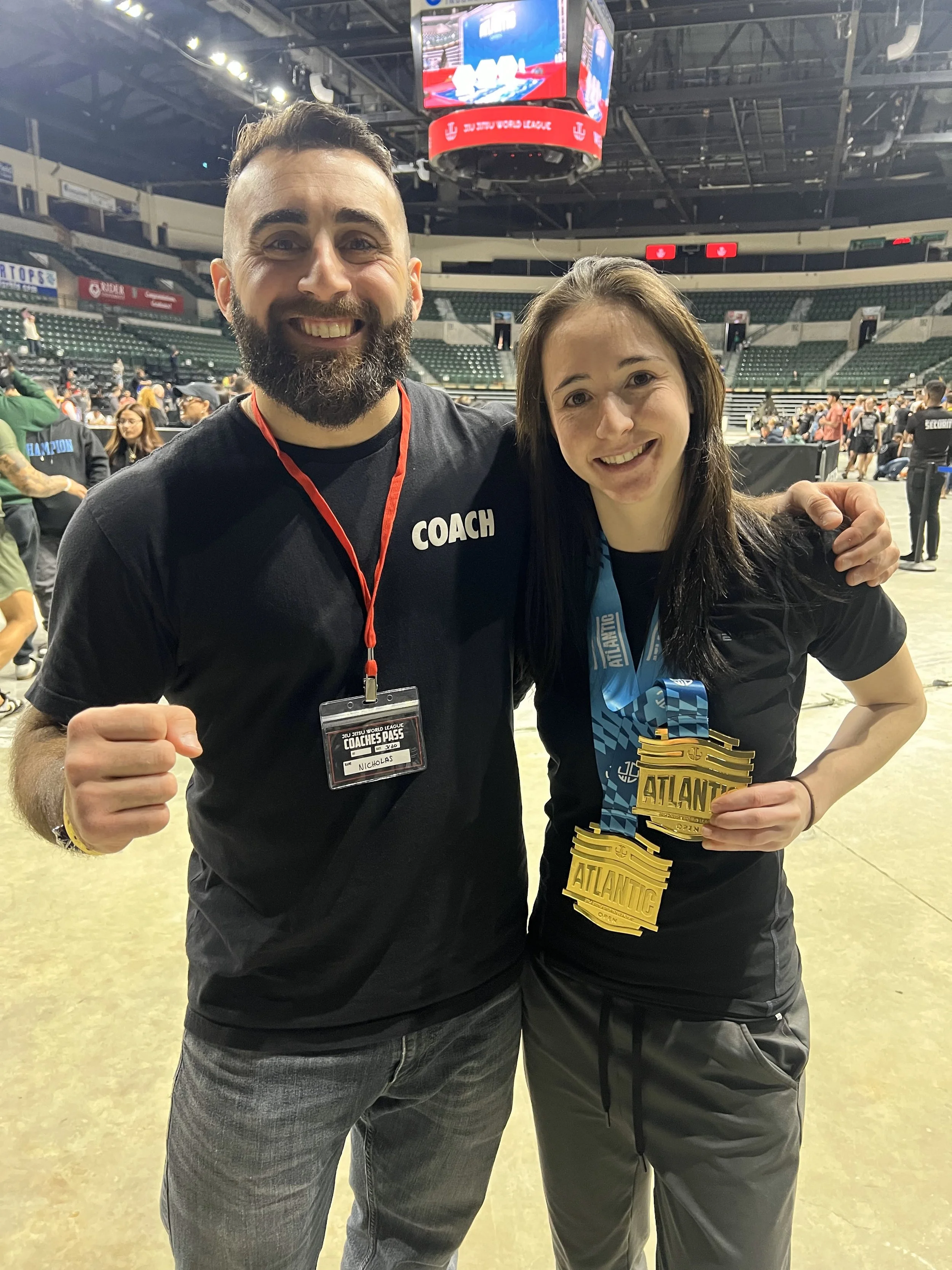 A man and woman smiling and posing together at a sports event in an indoor arena, with medals hanging from their necks, the woman holding her medals, and the man wearing a "COACH" shirt.