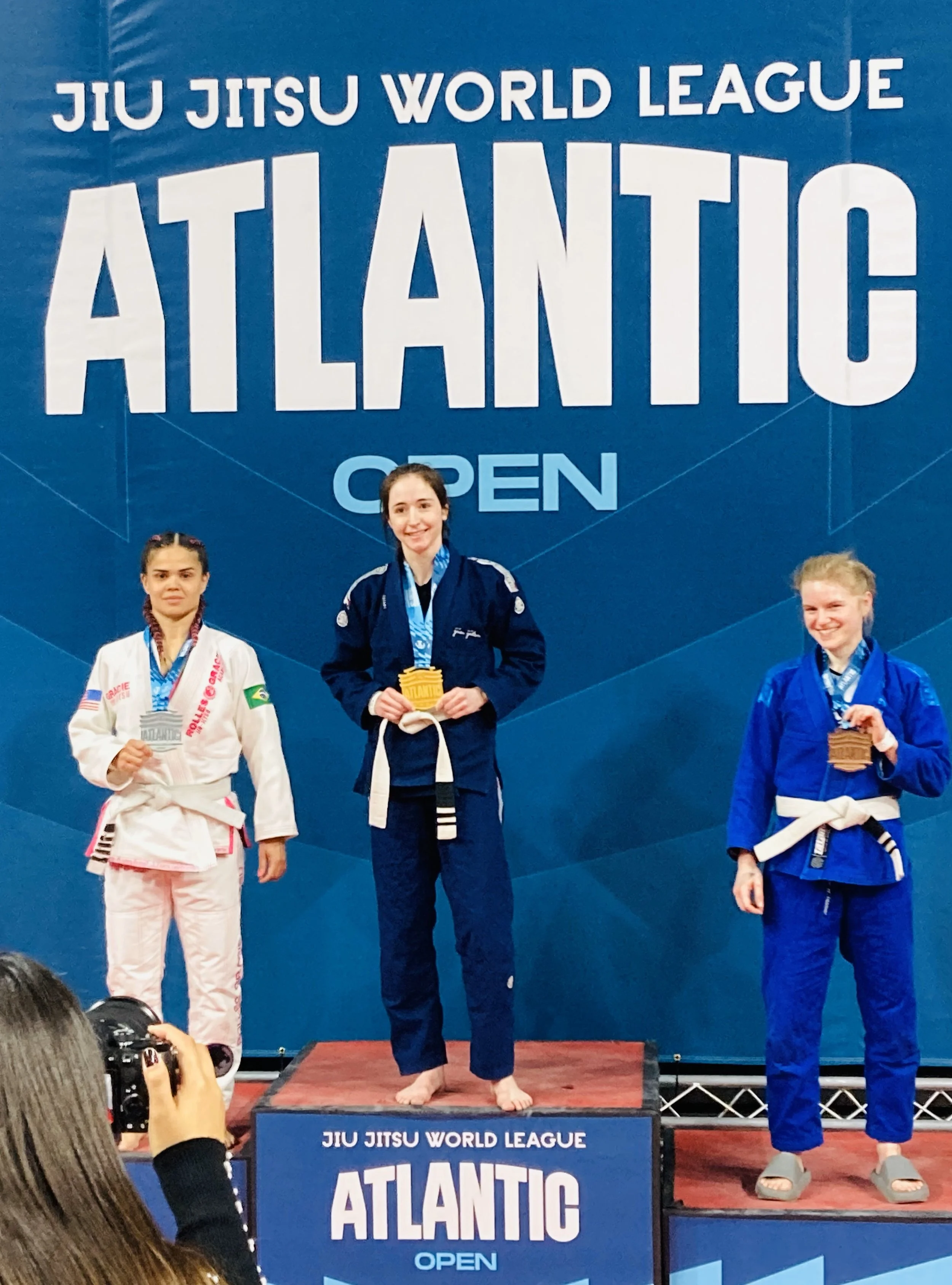 Three women in judo uniforms on a winners' podium at a jiu jitsu competition, holding medals, with a blue backdrop reading 'Jiu Jitsu World League Atlantic Open'.