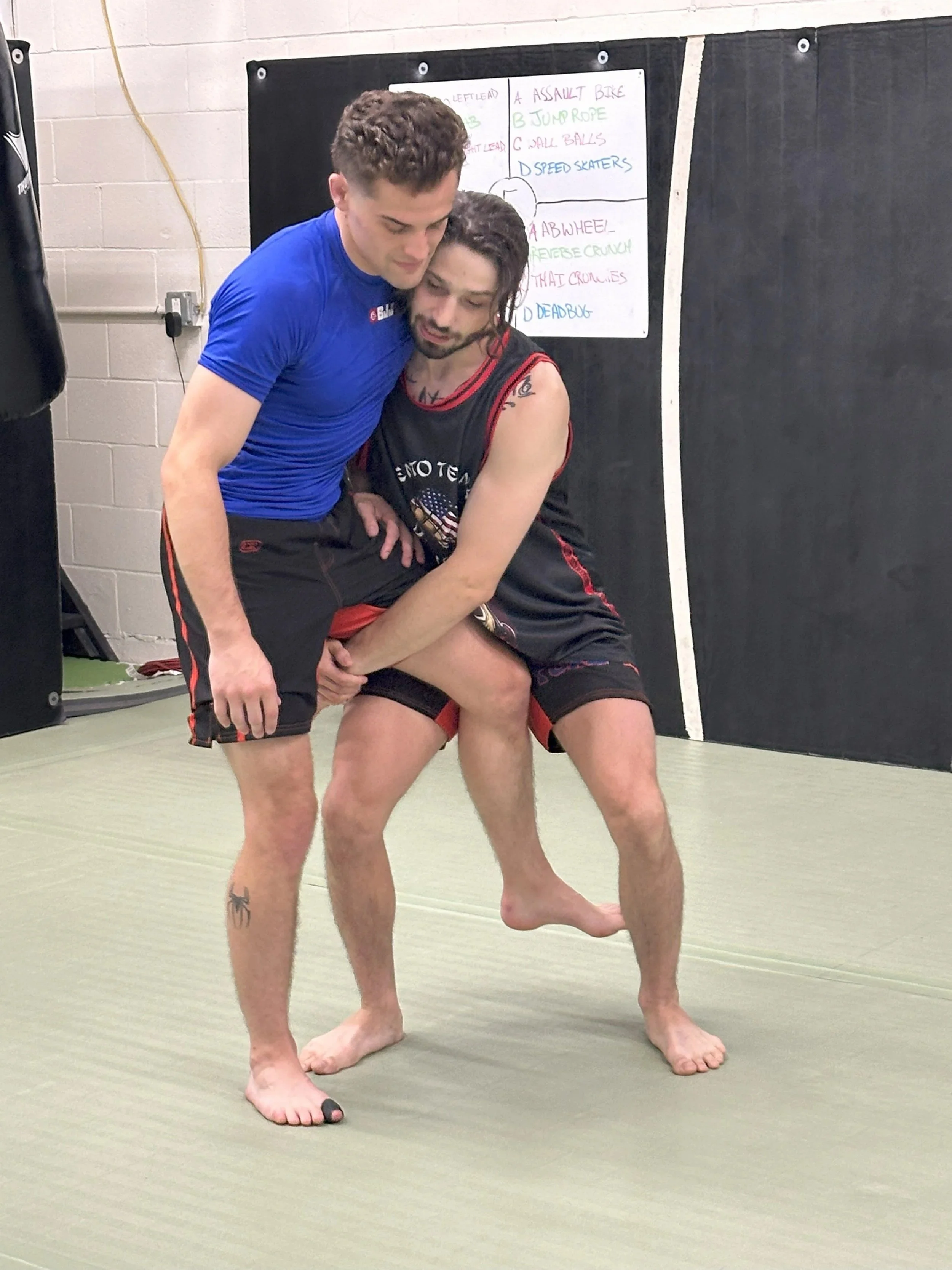Two men practicing Brazilian Jiu-Jitsu, with one man in a blue shirt and the other wearing a black gi, on a mat in a training room.