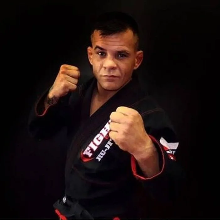 A man in a black Brazilian Jiu-Jitsu gi, making a fist with her right hand and posing in a fighting stance against a black background.