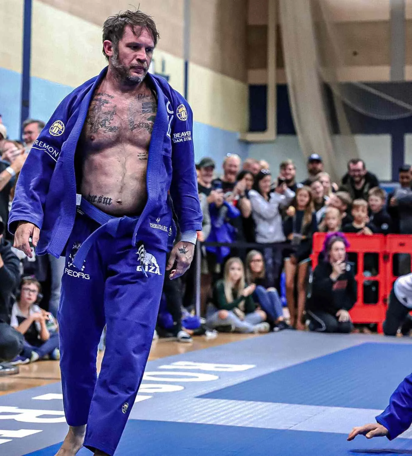 Actor Tom Hardy in a Brazilian Jiu-Jitsu competition in a blue gi walking on a mat, with a crowd of spectators watching in the background.