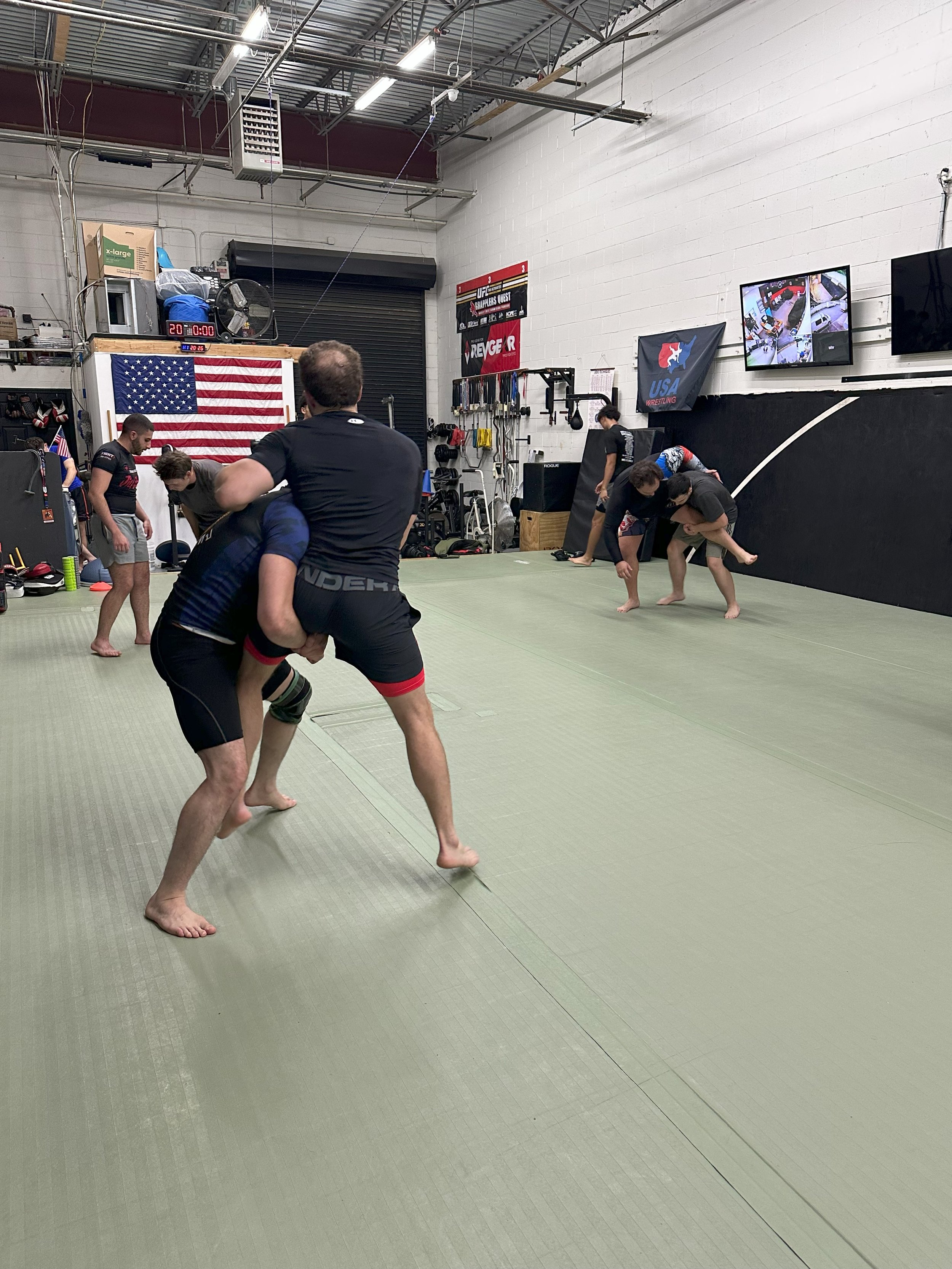 People practicing Brazilian Jiu-Jitsu in a martial arts gym with American flag and fitness equipment in the background.