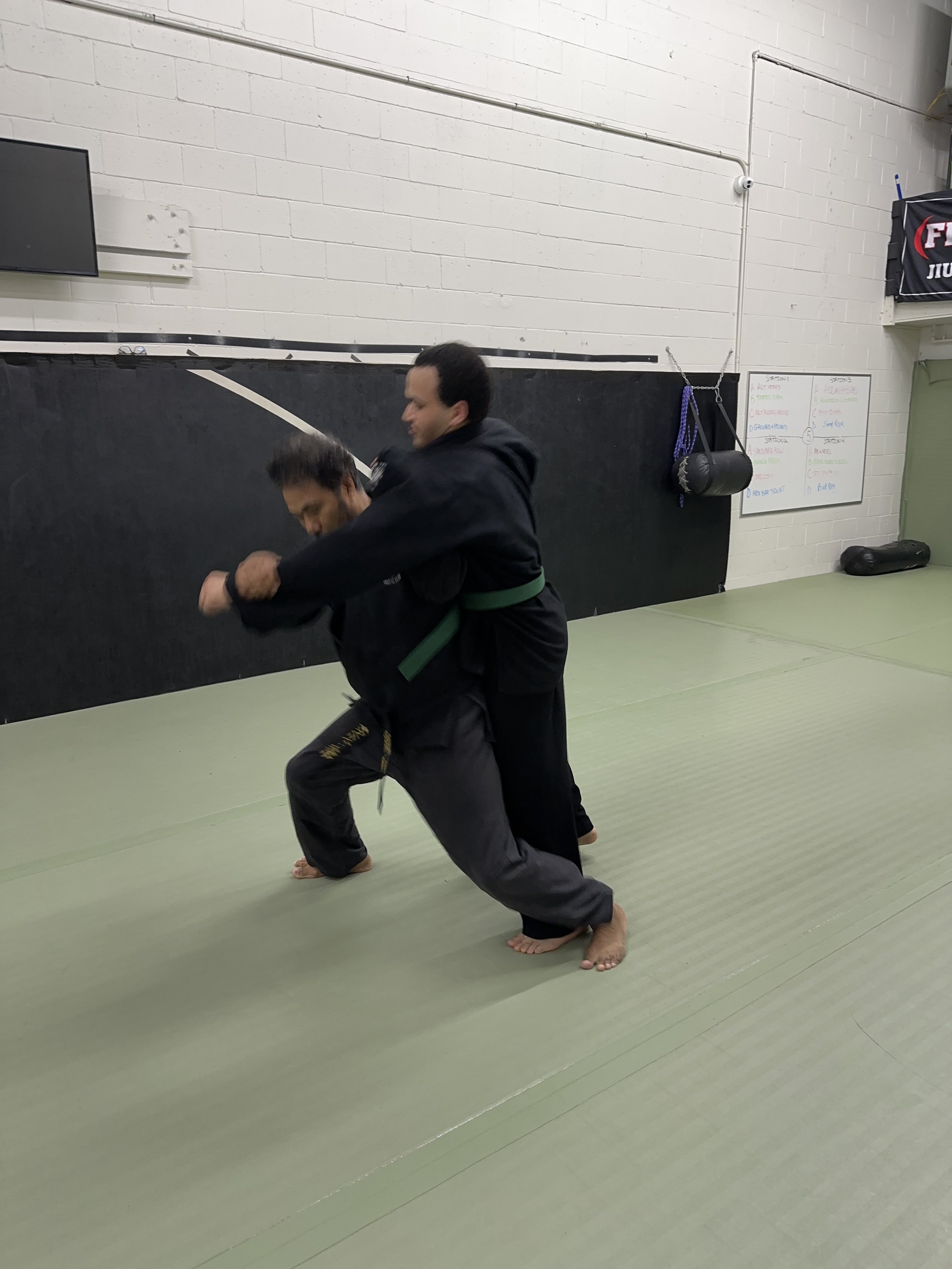 Two martial artists wearing black uniforms practicing judo with one applying a throw on the other in a martial arts gym.
