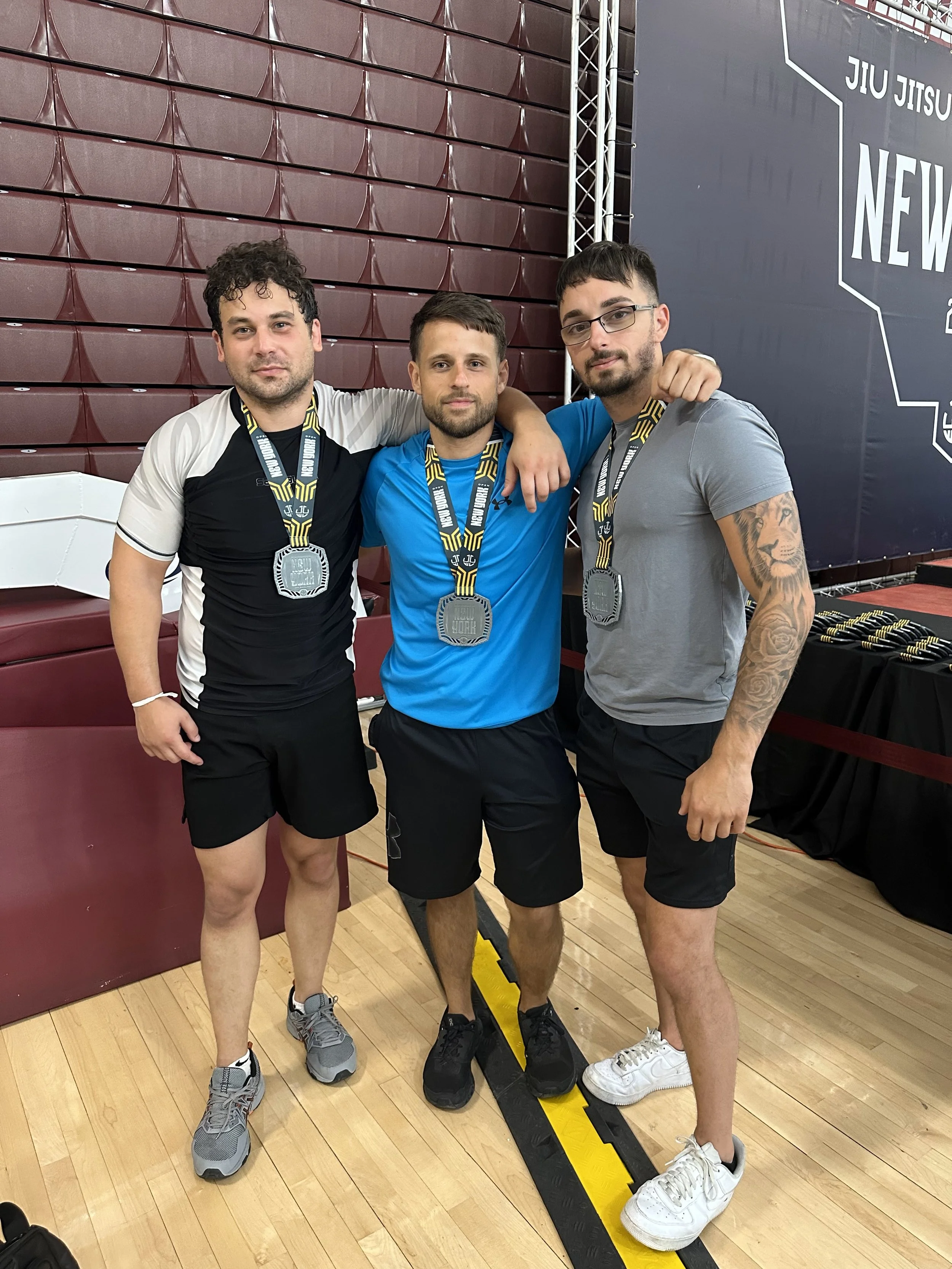 Three men posing with medals at a sports event, standing on a gymnasium floor, with a large banner reading 'New York' and 'Jiu Jitsu' behind them.