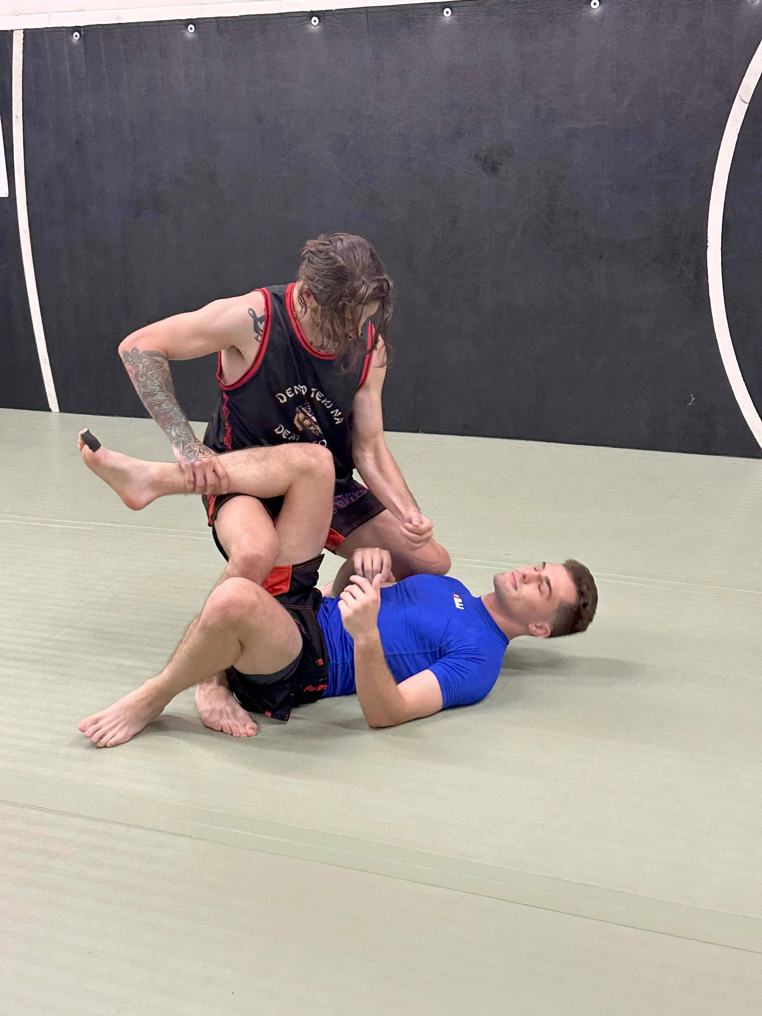 Two people practicing Brazilian jiu-jitsu on a mat in a gym, one person in a black gi with tattoos and the other in a blue shirt, engaged in a grappling position.