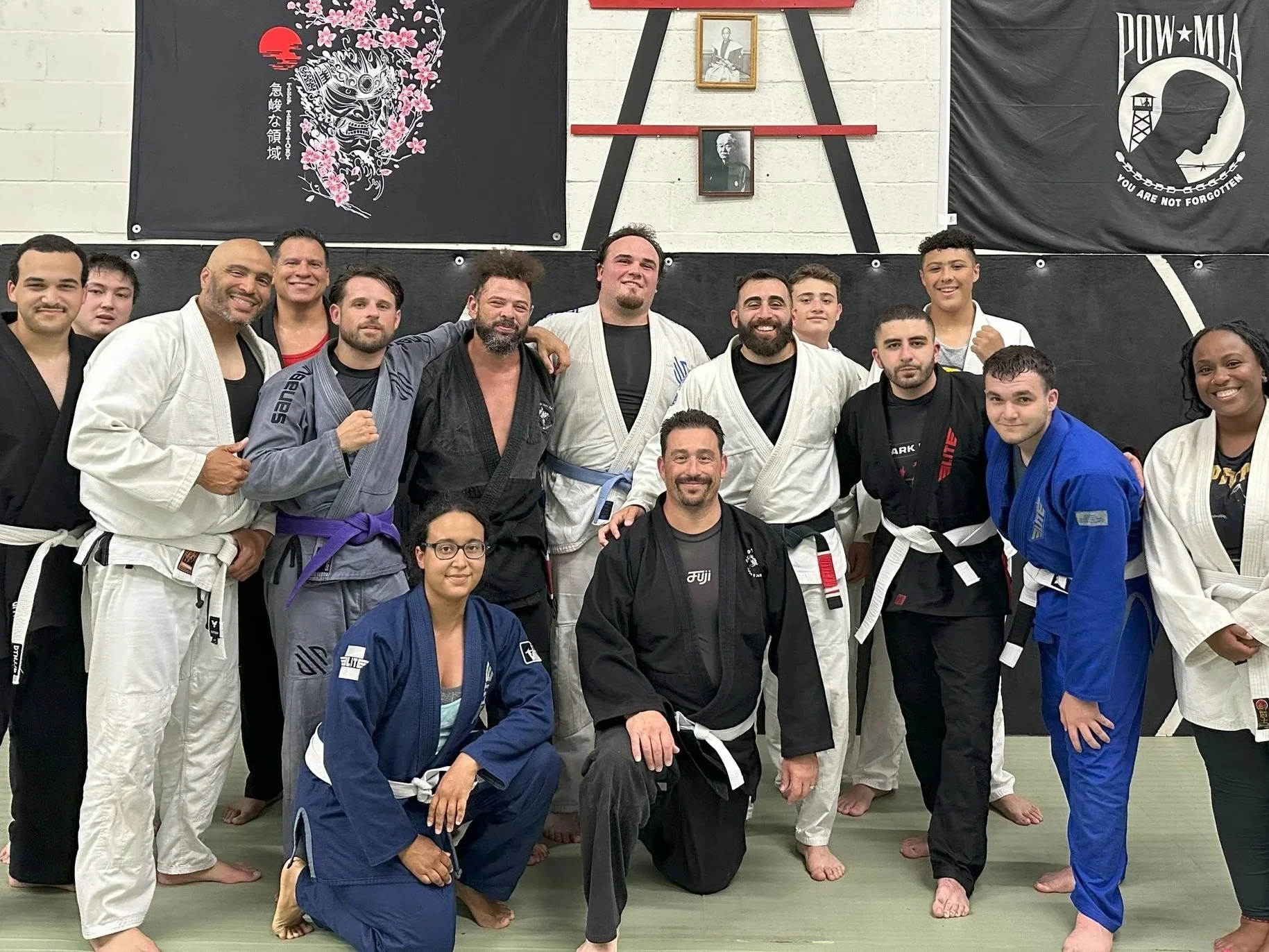 Group of Brazilian Jiu-Jitsu practitioners in gi uniforms posing for a photo in a dojo, with banners and portraits on the wall behind them.