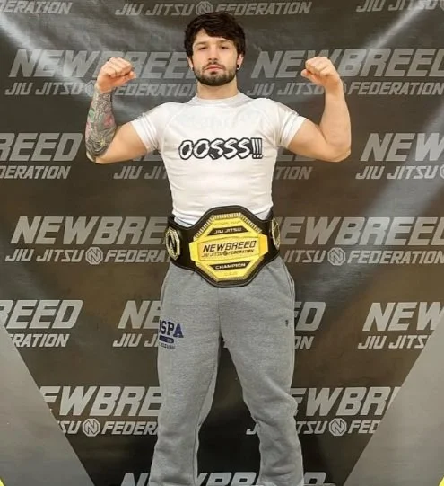 A young man with dark hair and facial hair, flexing his biceps in a gym or training center. He is wearing a white T-shirt that says 'OOSSS!!!' and grey sweatpants. He has a tattoo on his left arm. He is standing in front of a black backdrop with repeating logos for 'NEW BREED' and 'Jiu Jitsu Federation,' and he is wearing a yellow championship belt around his waist that says 'NEW BREED'.