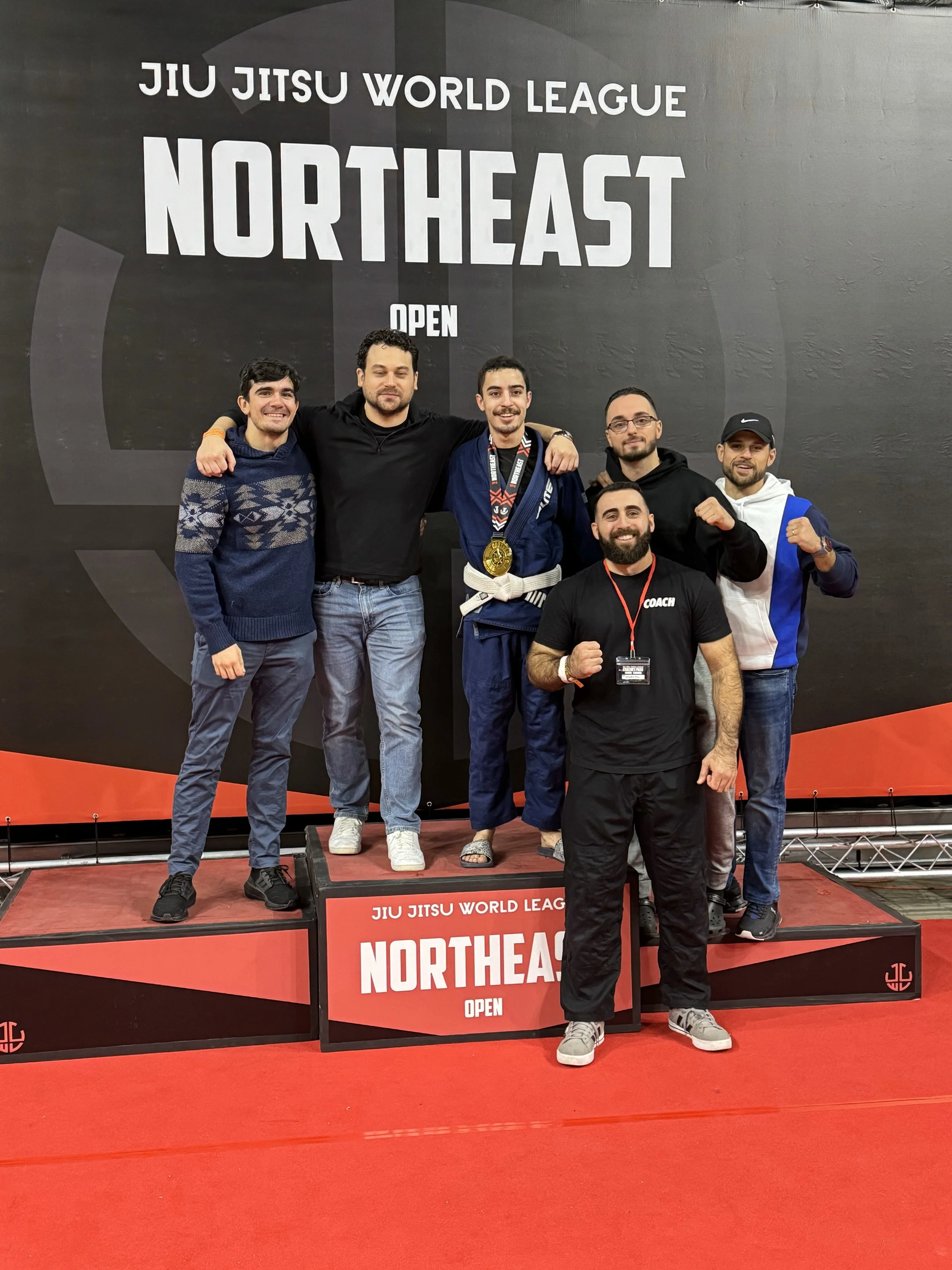 Six men posing on a winners' podium at a jiu jitsu competition, with a large sign reading 'Jiu Jitsu World League Northeast Open' behind them. One man in the center wears a blue gi with medals around his neck.