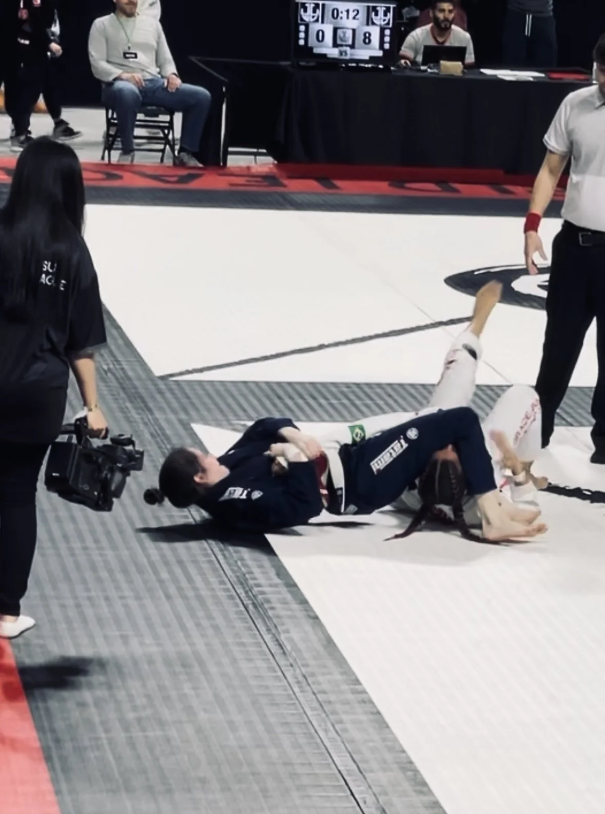 Two female Brazilian Jiu-Jitsu competitors on the mat, one is on her back and the other is on top, during a match at a tournament. An effective demonstration of an arm bar, a technique which attacks the elbow joint, is being applied in a competition.