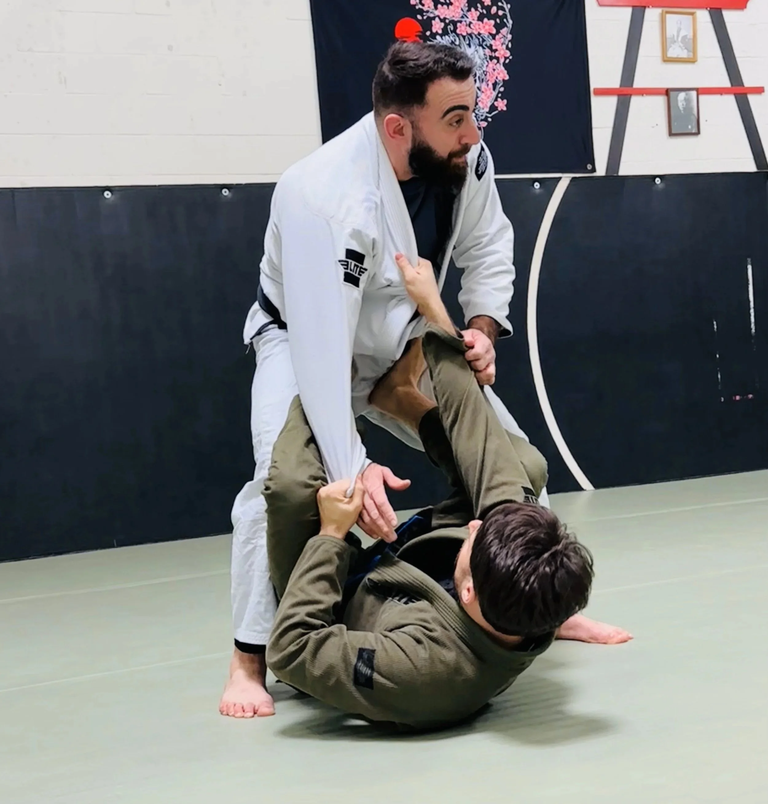 Two men practicing Brazilian Jiu-Jitsu on a mat; one man wearing a white gi is standing and controlling the other man in an olive gi who is on the ground, in an arm control position.