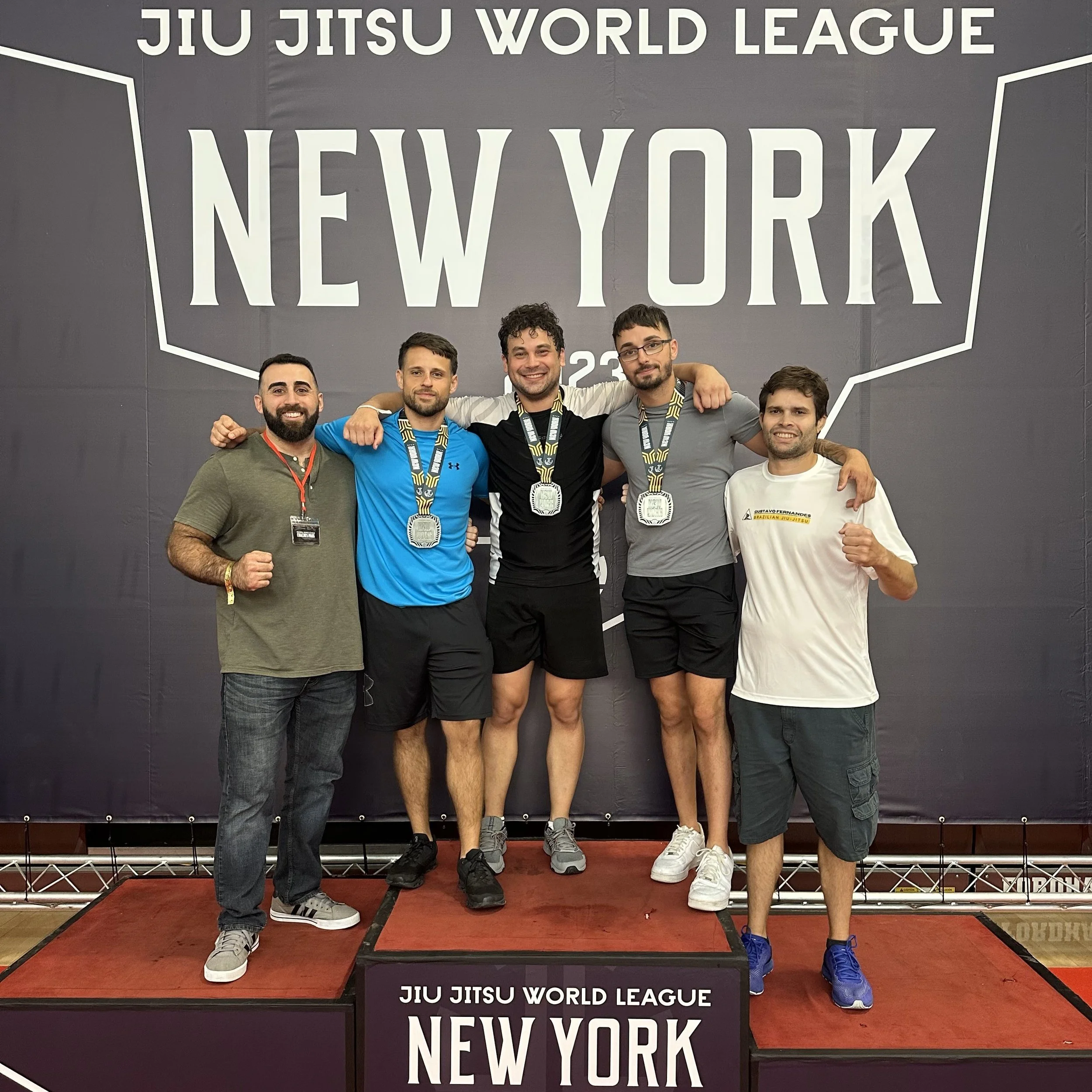 Group of five men standing on a winners' podium at a Jiu Jitsu competition in New York, wearing medals and smiling.