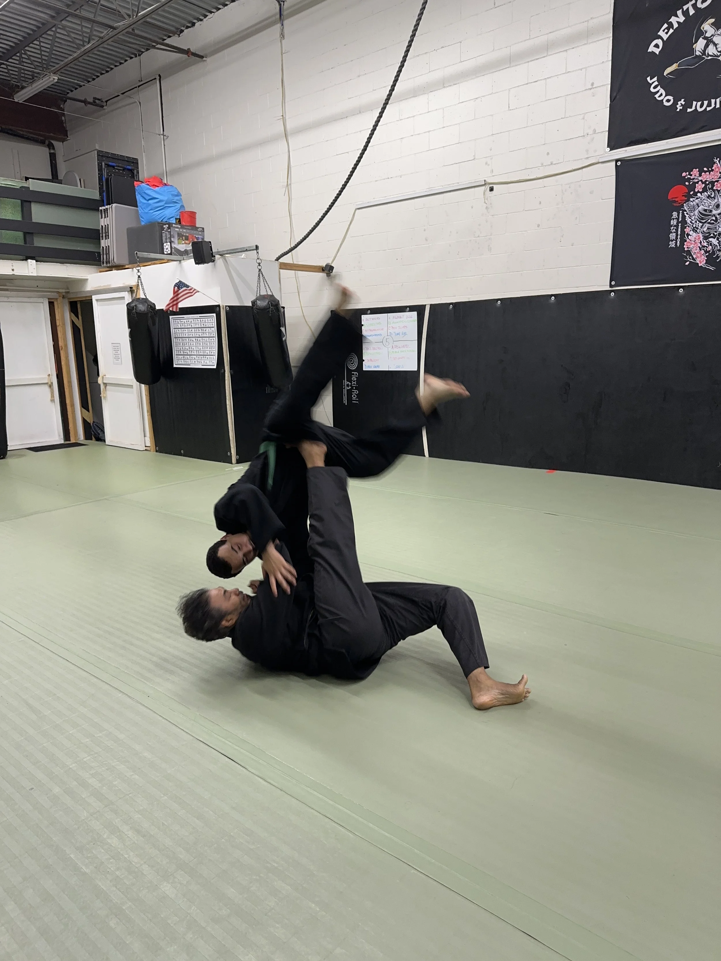 Two individuals practicing judo on a green mat, one on the ground and the other attempting a move, in a martial arts gym.