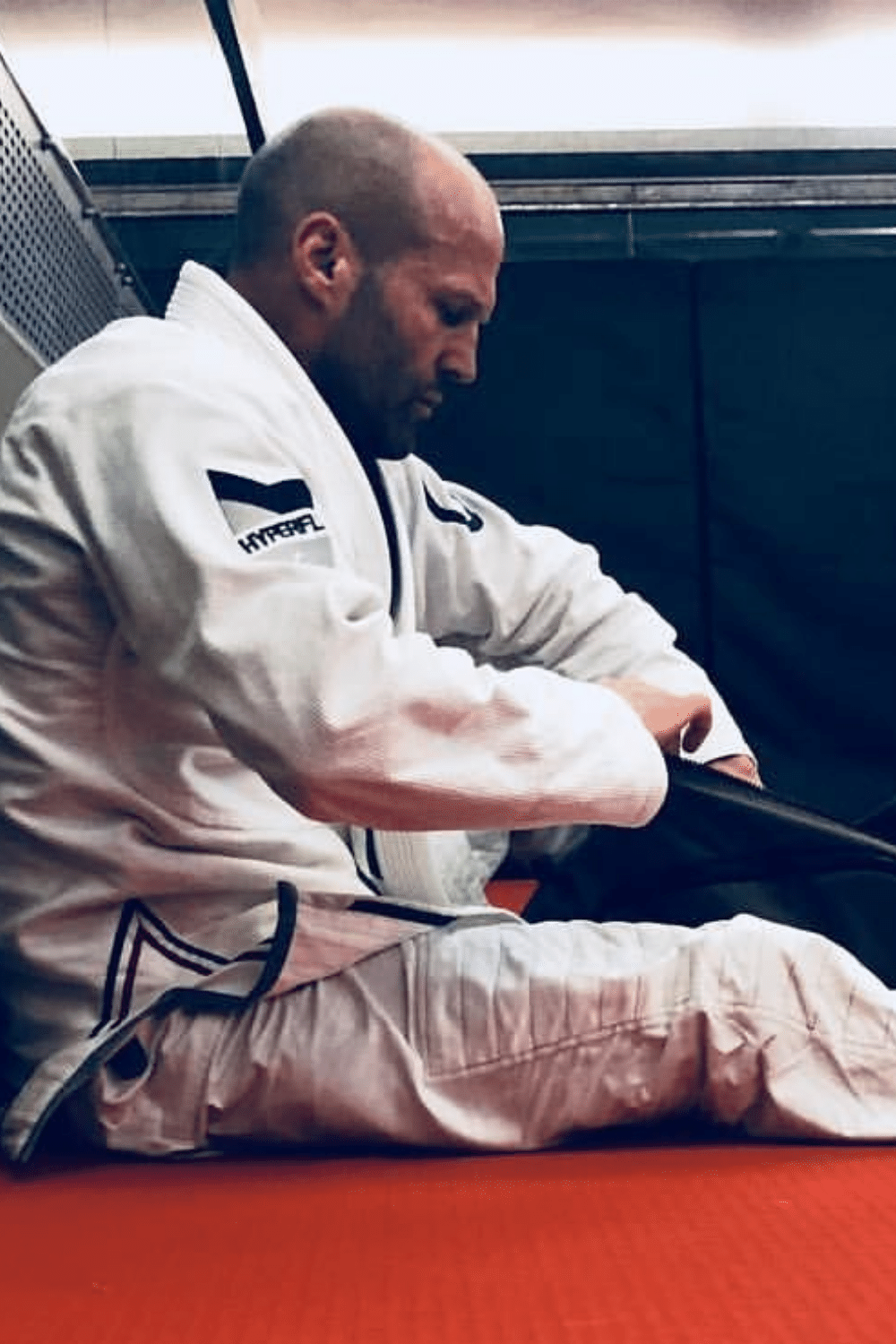 Actor Jason Statham in a white martial arts gi sitting on a red mat, adjusting his black belt, with a focused expression.