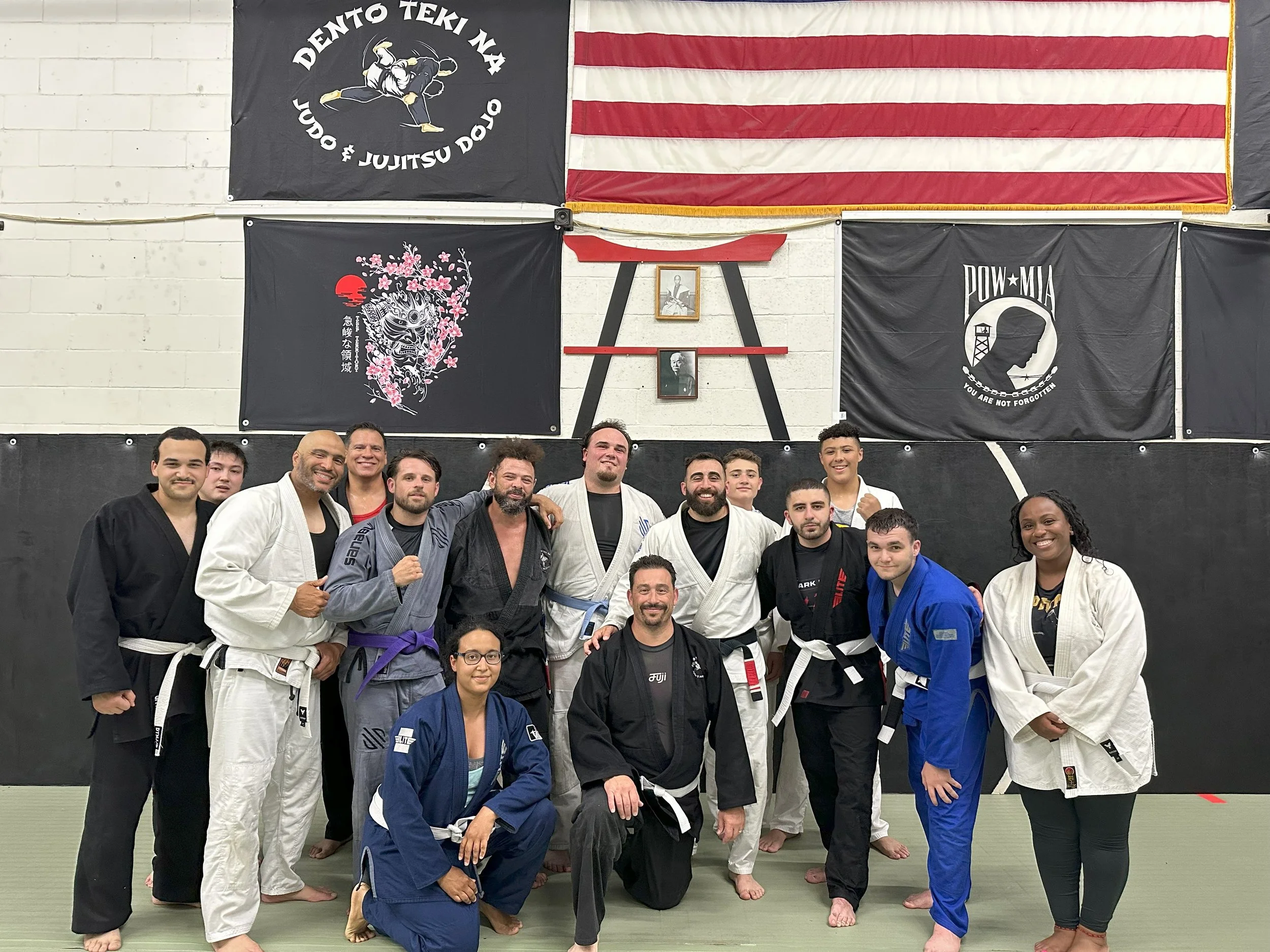 Group of people in Brazilian Jiu-Jitsu gis and uniforms posing for a photo inside a gym with flags and banners on the wall, including an American flag and flags with martial arts logos.