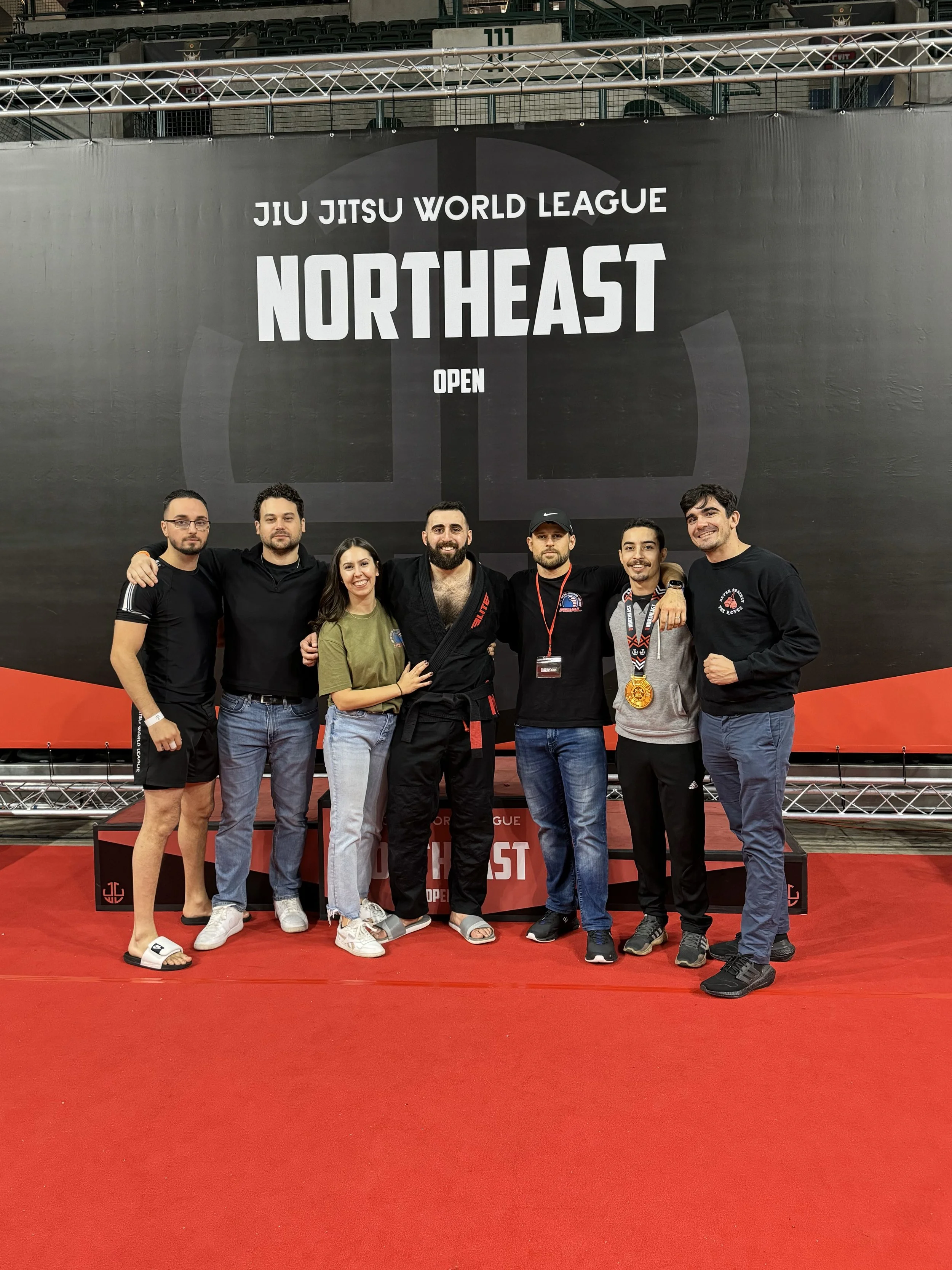 Group of seven people standing in front of a large sign that reads 'Jiu Jitsu World League Northeast Open' at a martial arts competition, with a red carpeted floor.