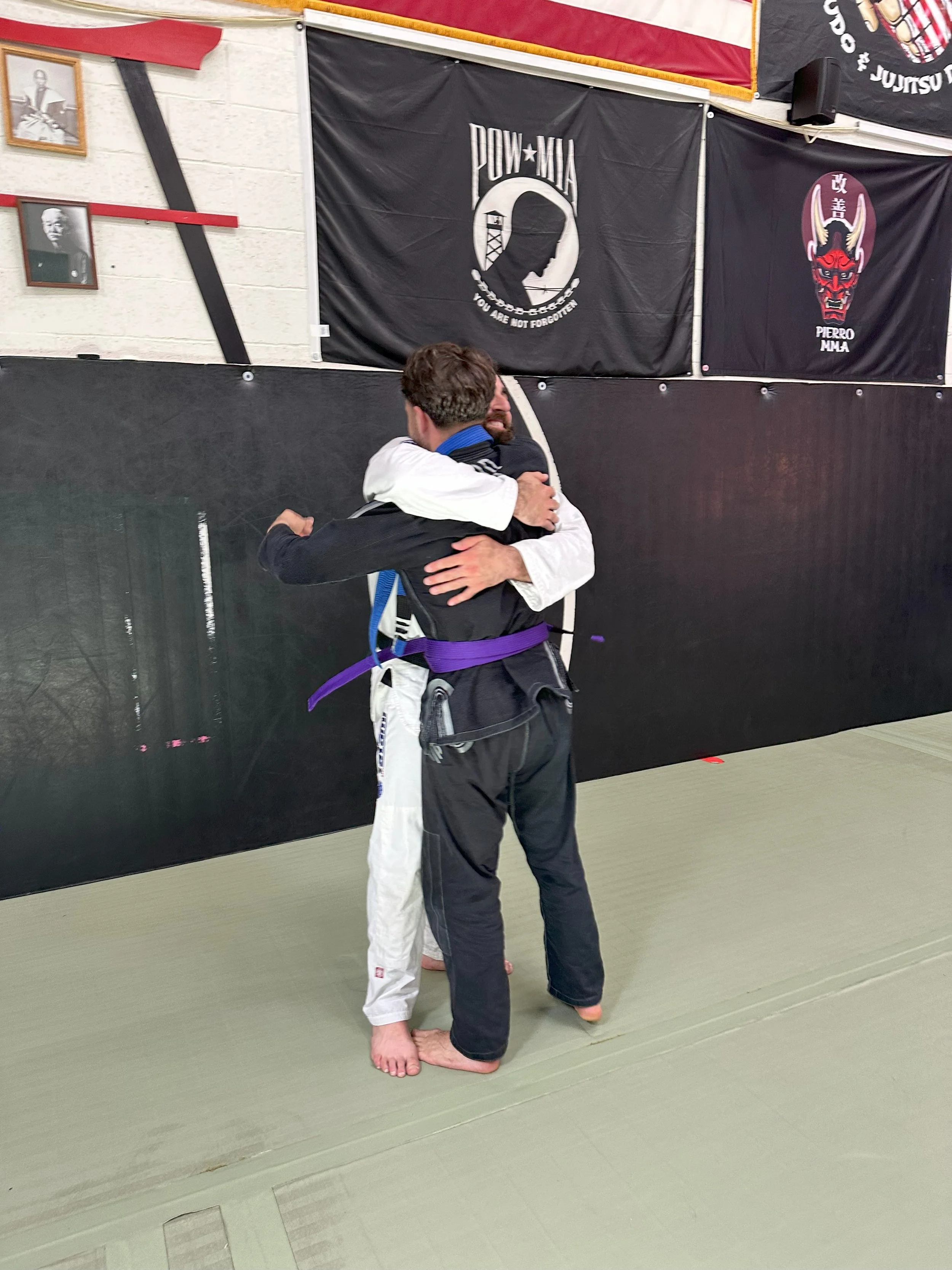 Two men hugging after a belt promotion in a Brazilian Jiu-Jitsu gym, where one student received their purple belt.