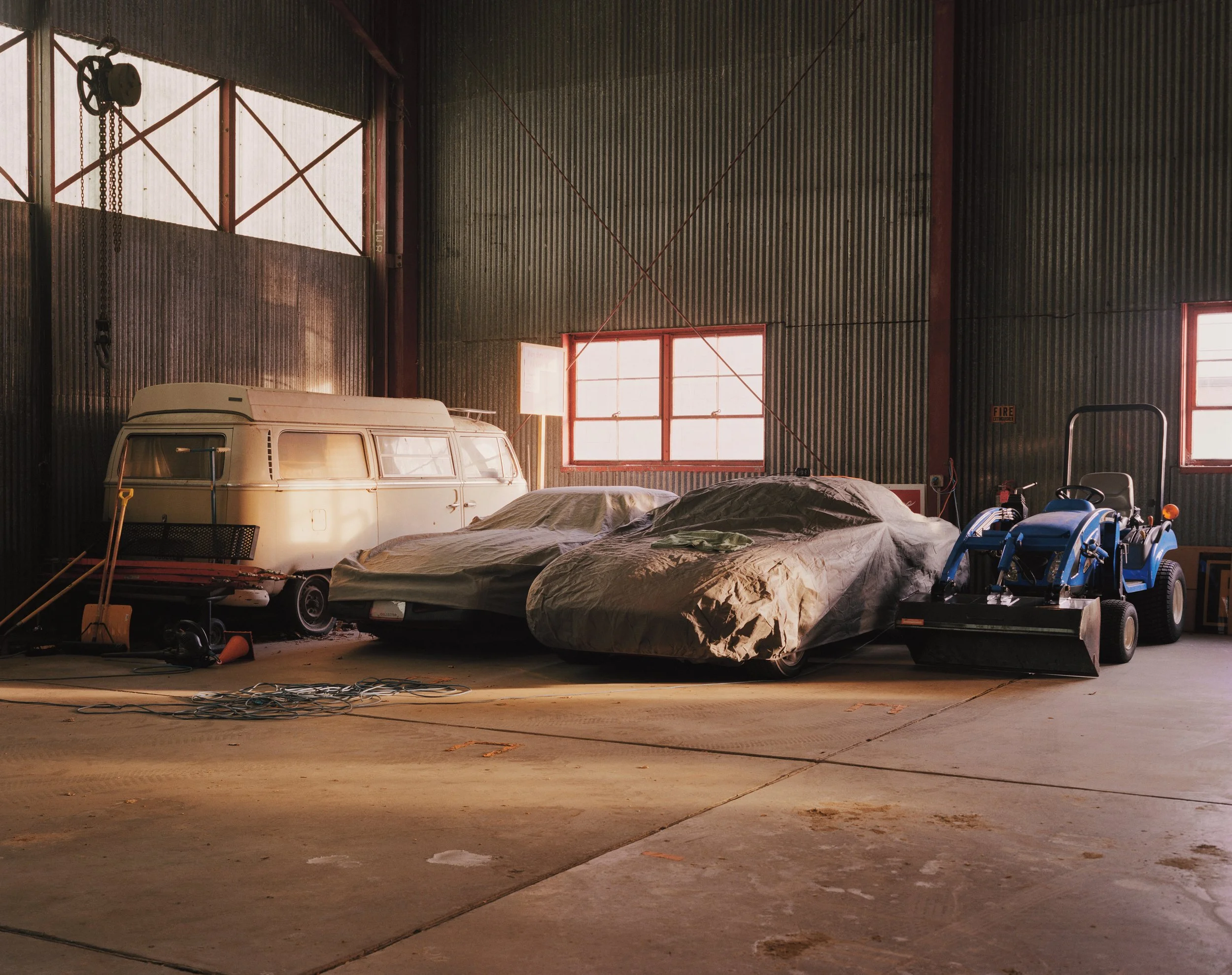 Camioneta blanca, autos cubiertos y un pequeño tractor azul en espacio interior contra una pared de aluminio